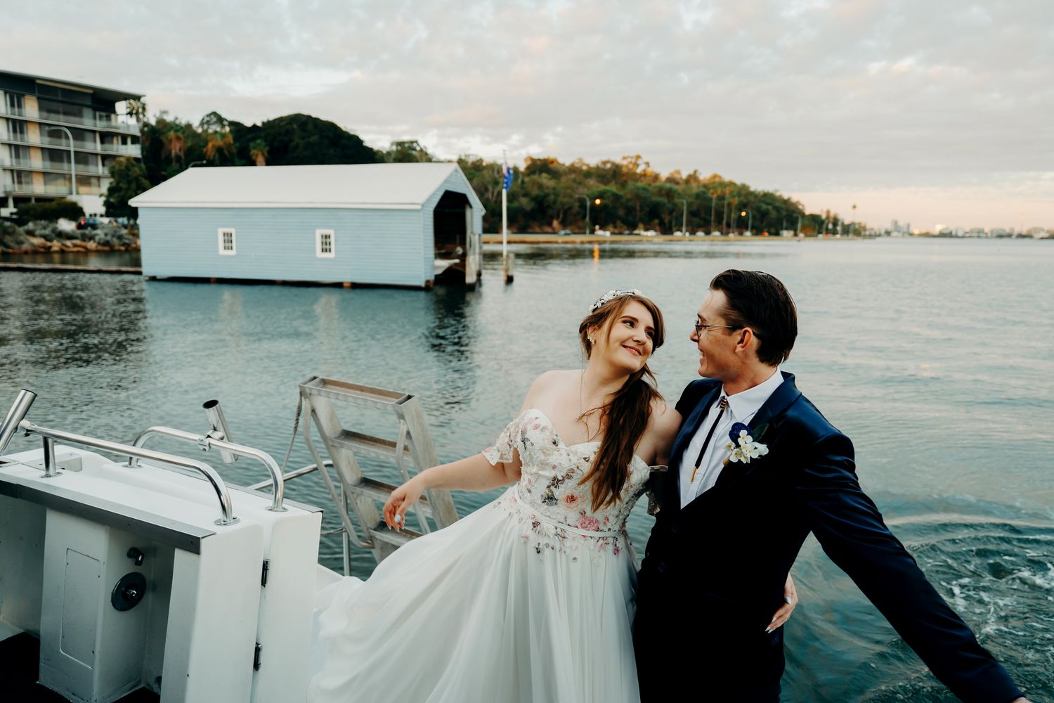 Happy couple celebrates on a boat with white boathouse and waterfront scenery at sunset.