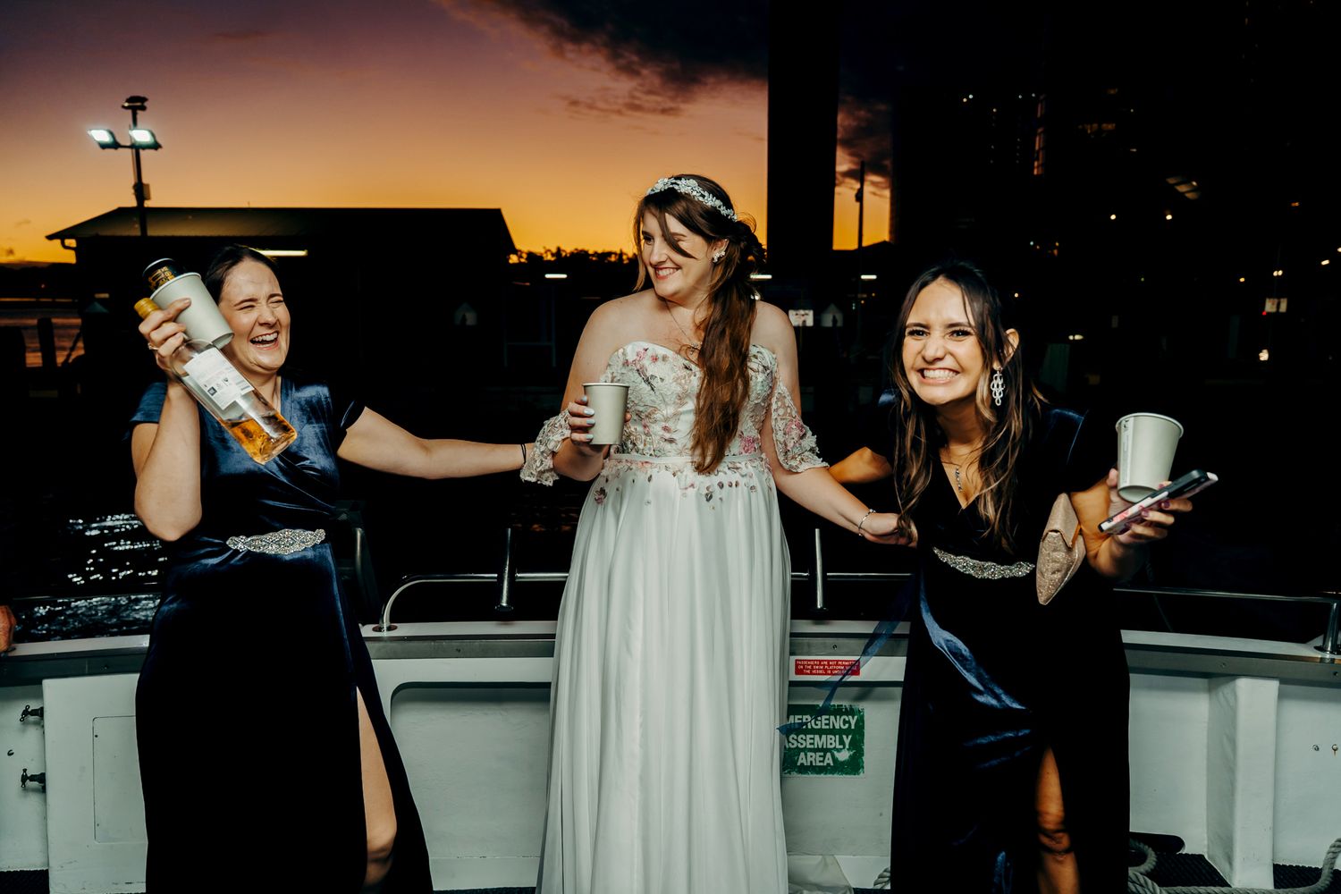 Three friends enjoying drinks and laughing together on a rooftop balcony during sunset.