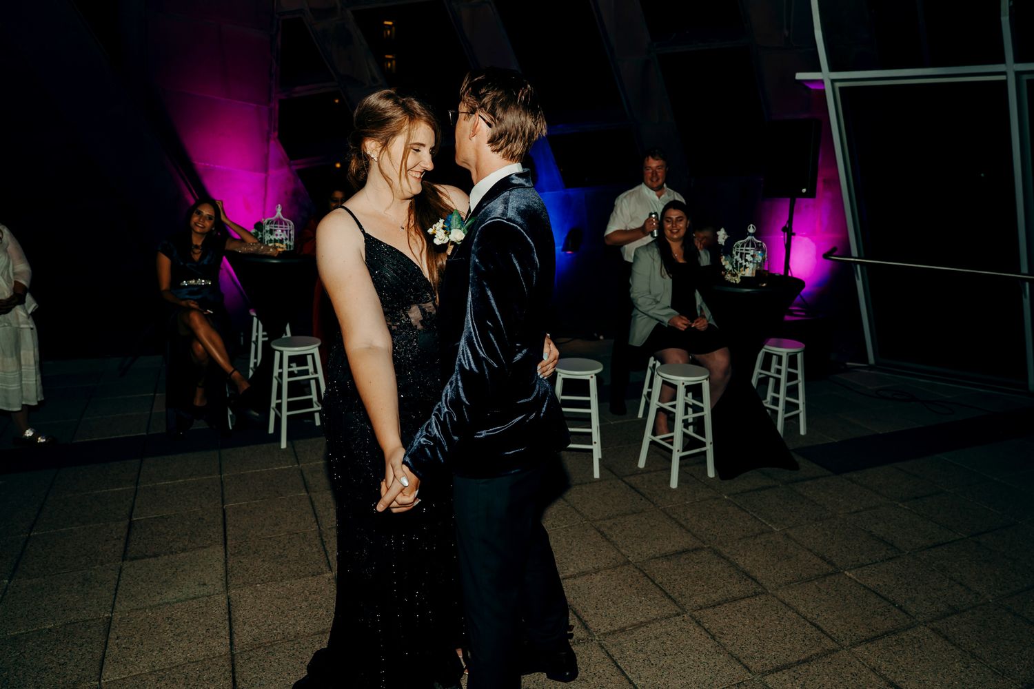 A couple shares an intimate dance moment at a reception with pink and blue mood lighting and bar stools in the background.