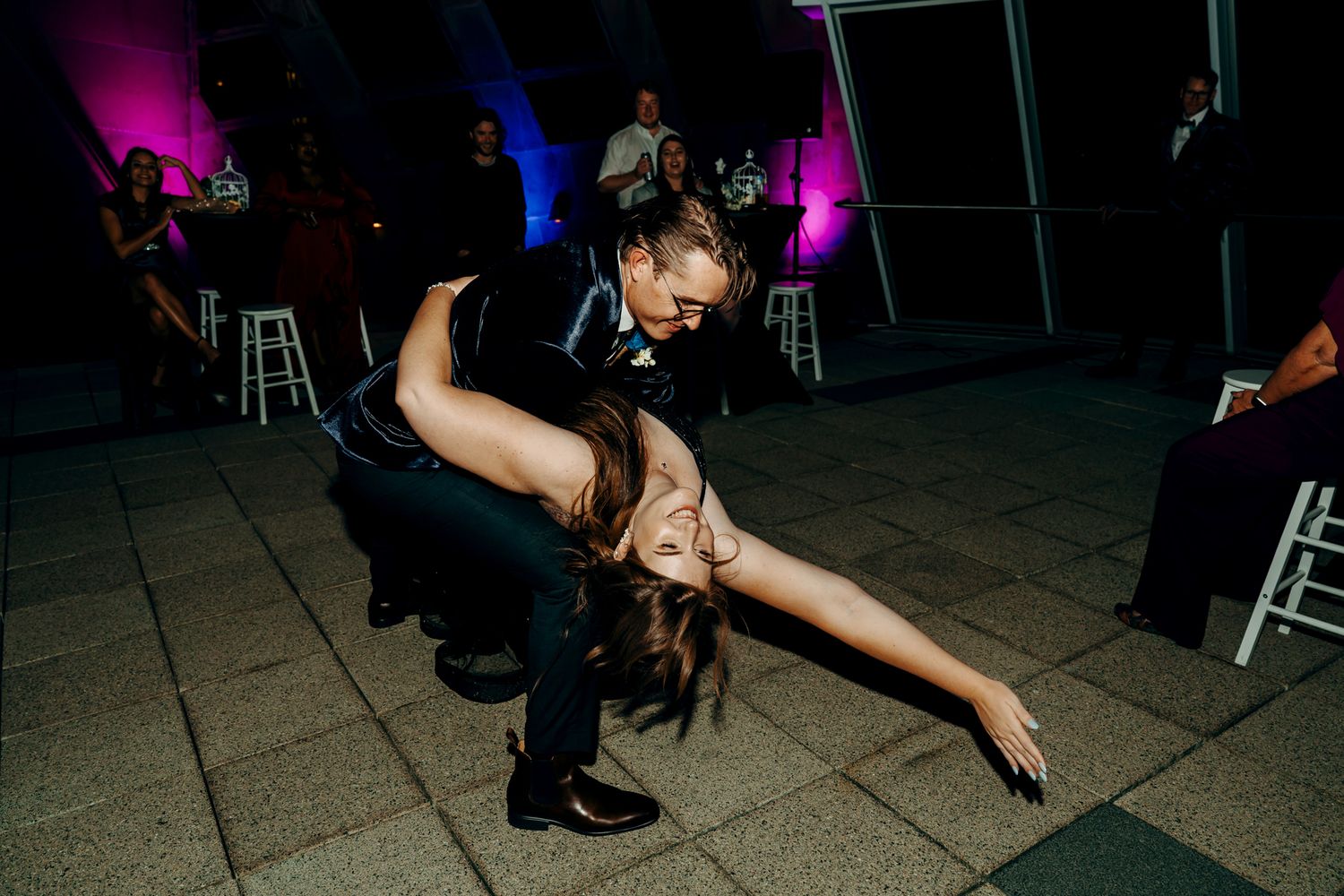 Party guests create fun dance moves on illuminated floor at nightclub with colorful purple and blue lighting.