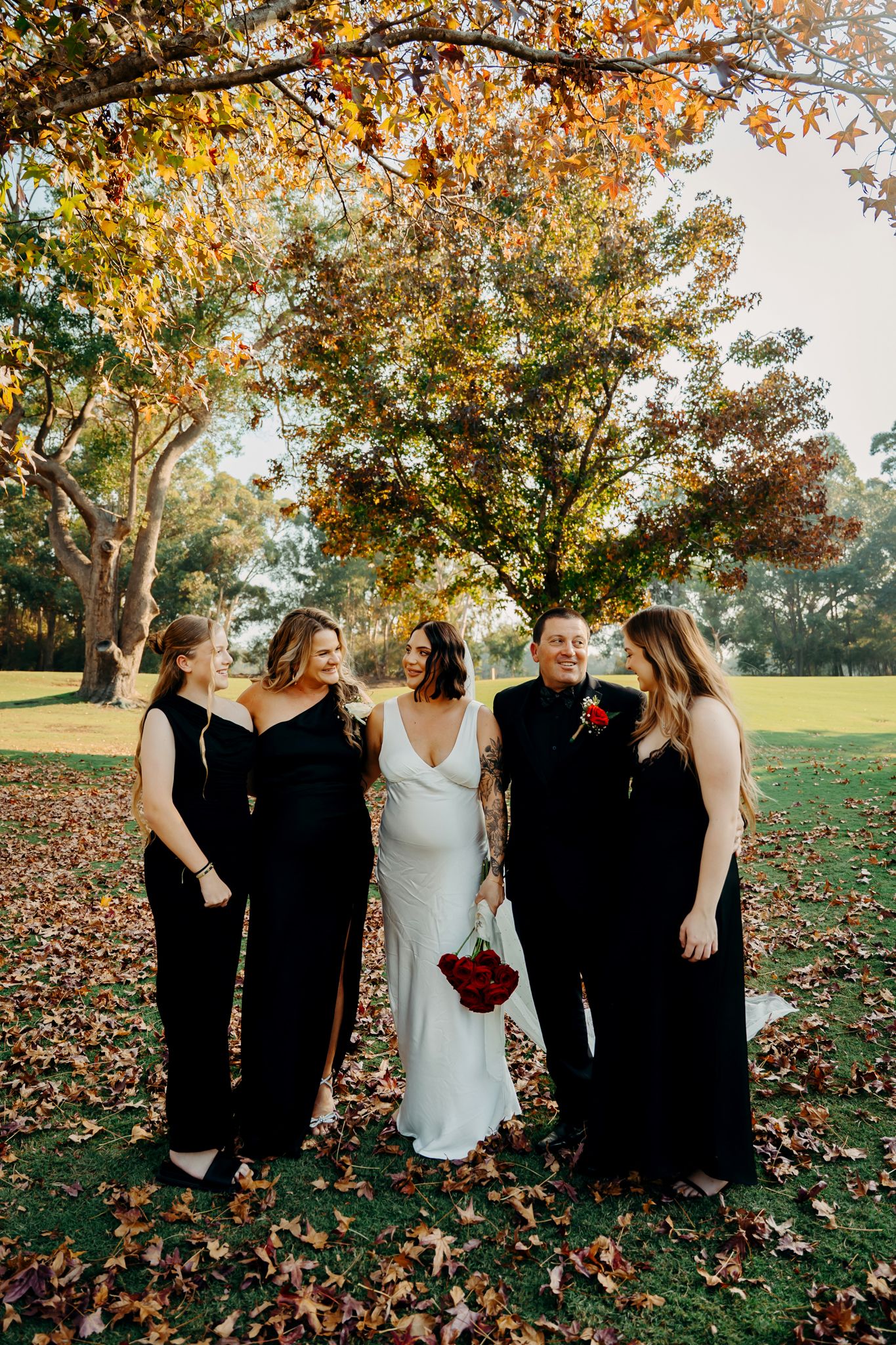 A wedding party stands together on grass covered with orange autumn leaves under golden fall foliage trees.