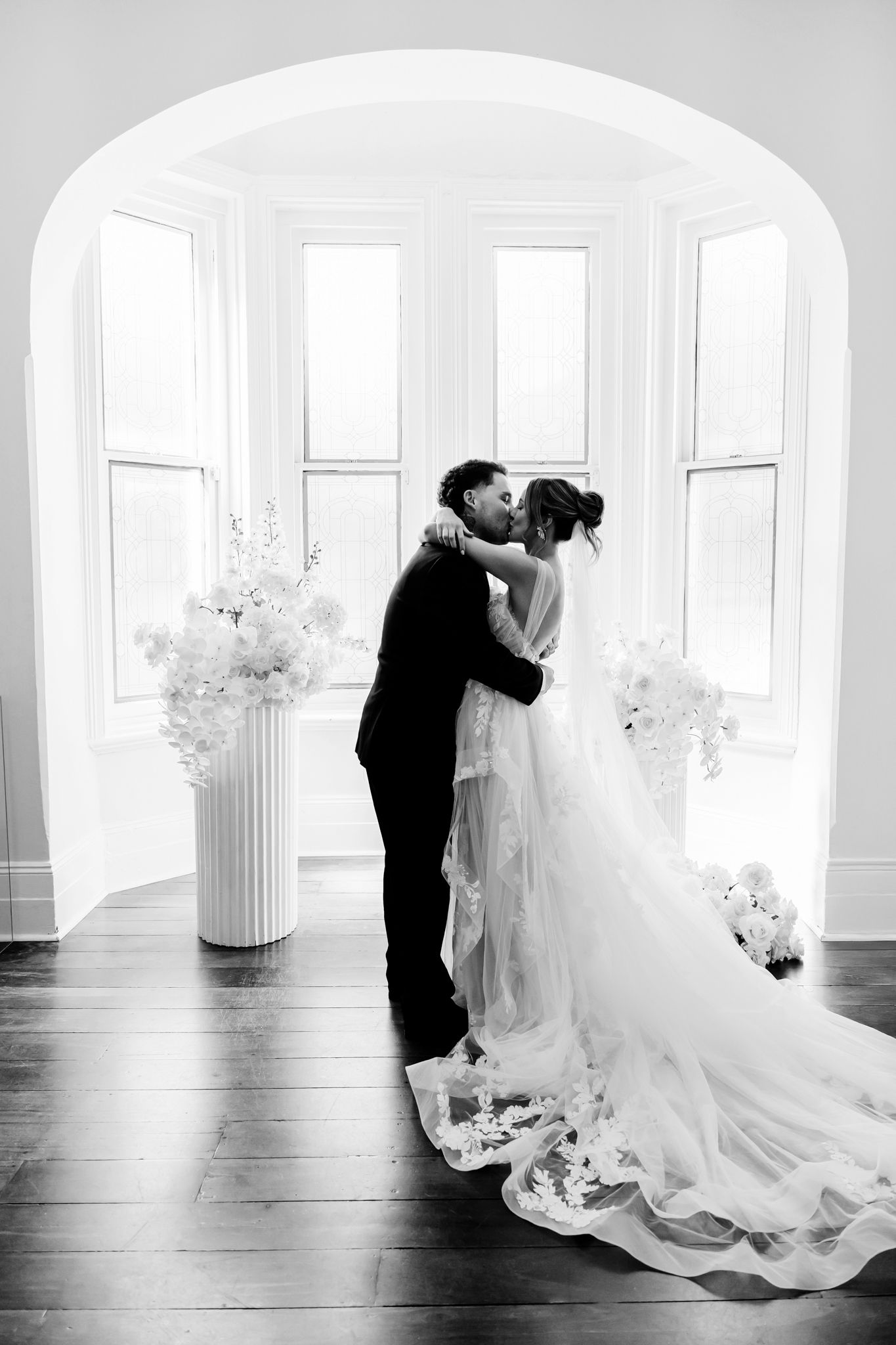 A romantic black and white wedding photo shows a couple embracing by bright windows with white floral arrangements.