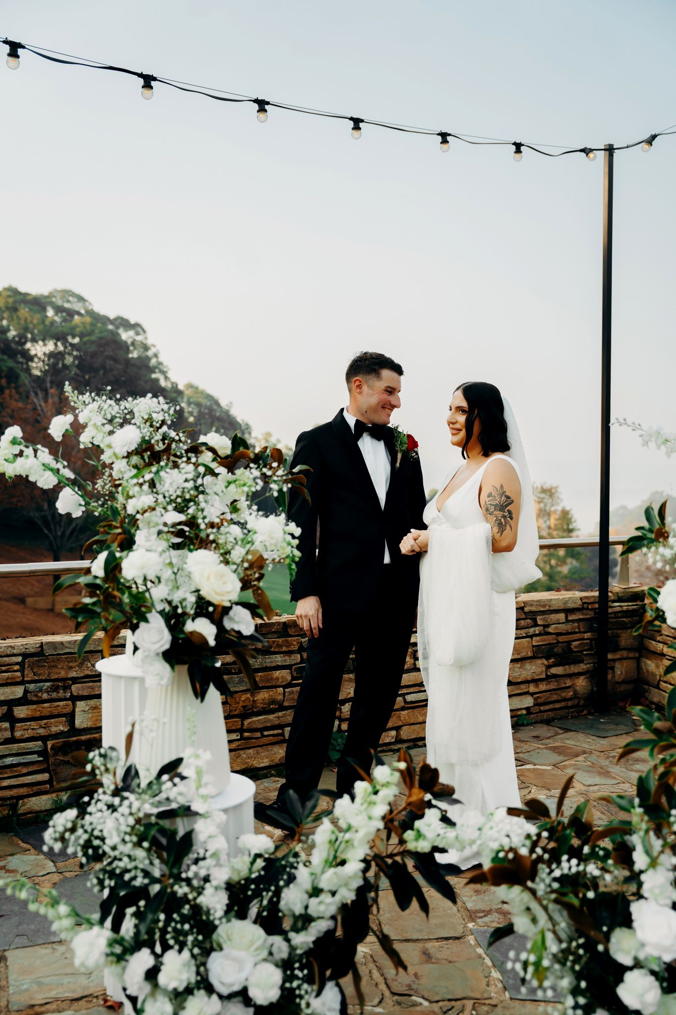 An elegant outdoor wedding ceremony setup with white floral arrangements and string lights on a stone patio.