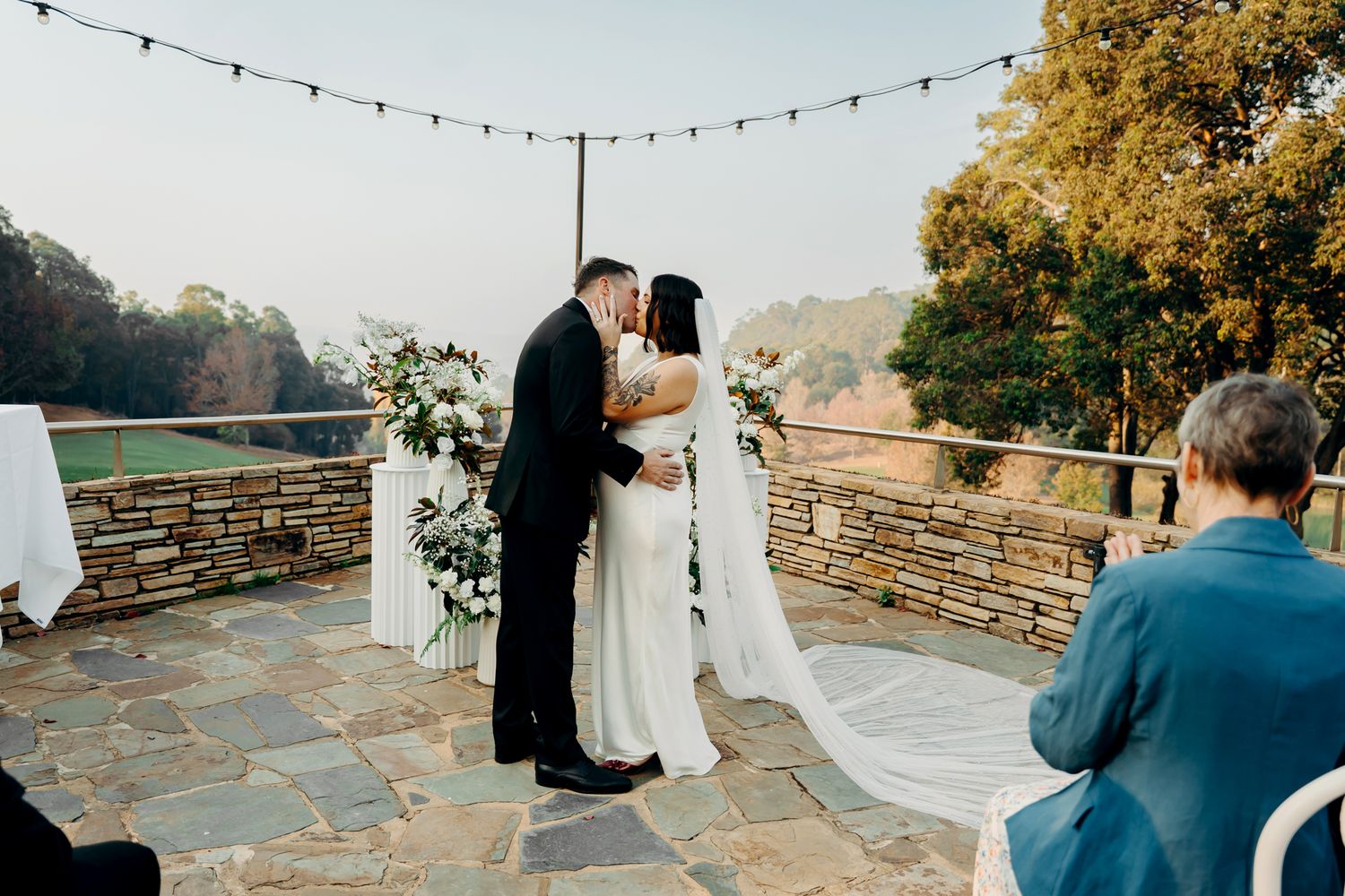 A string of twinkling lights hangs above a rustic stone wall terrace overlooking a scenic mountain landscape at sunset.