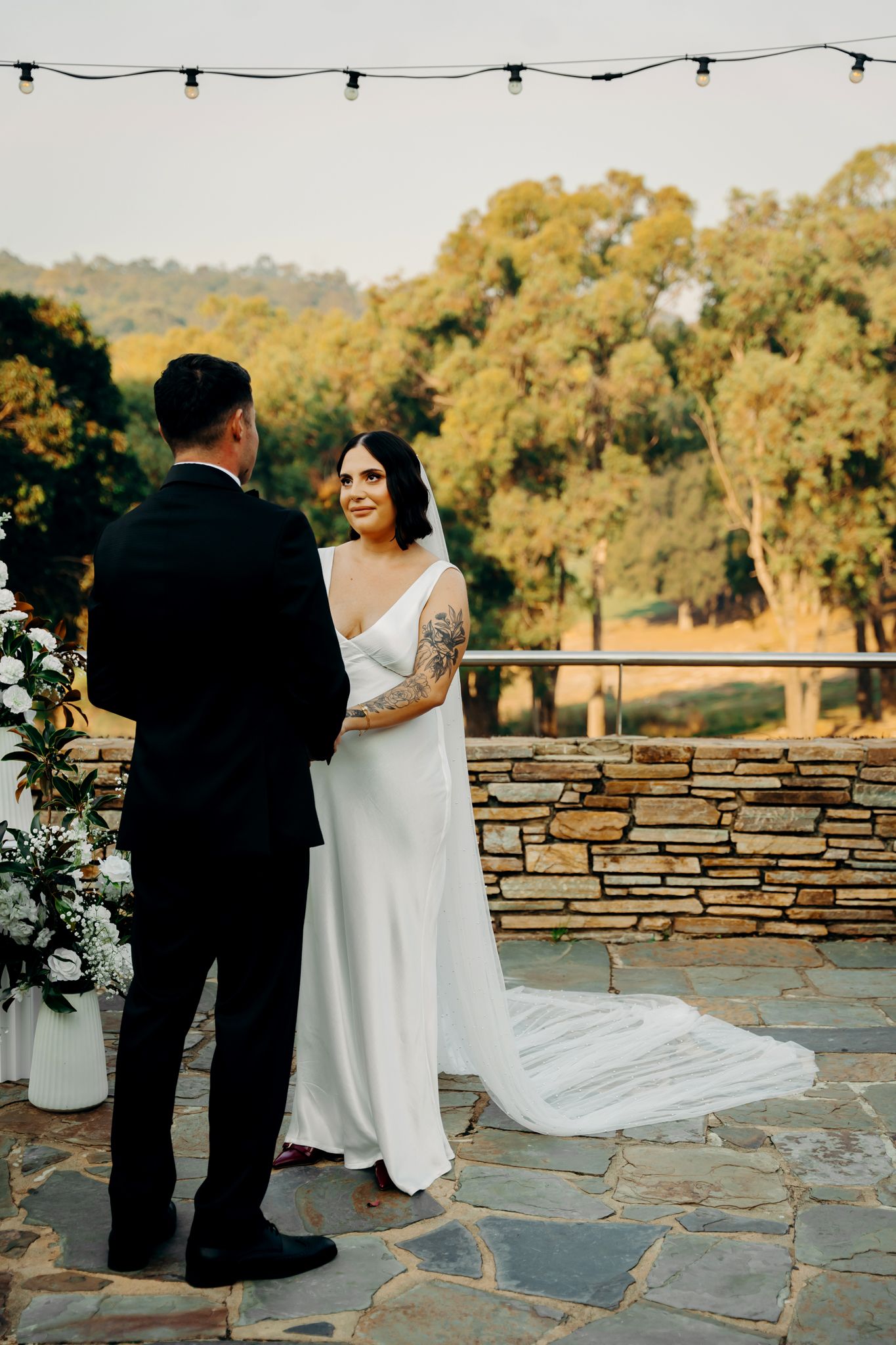 A bride and groom stand together at an outdoor wedding ceremony with stone wall and string lights during golden hour.
