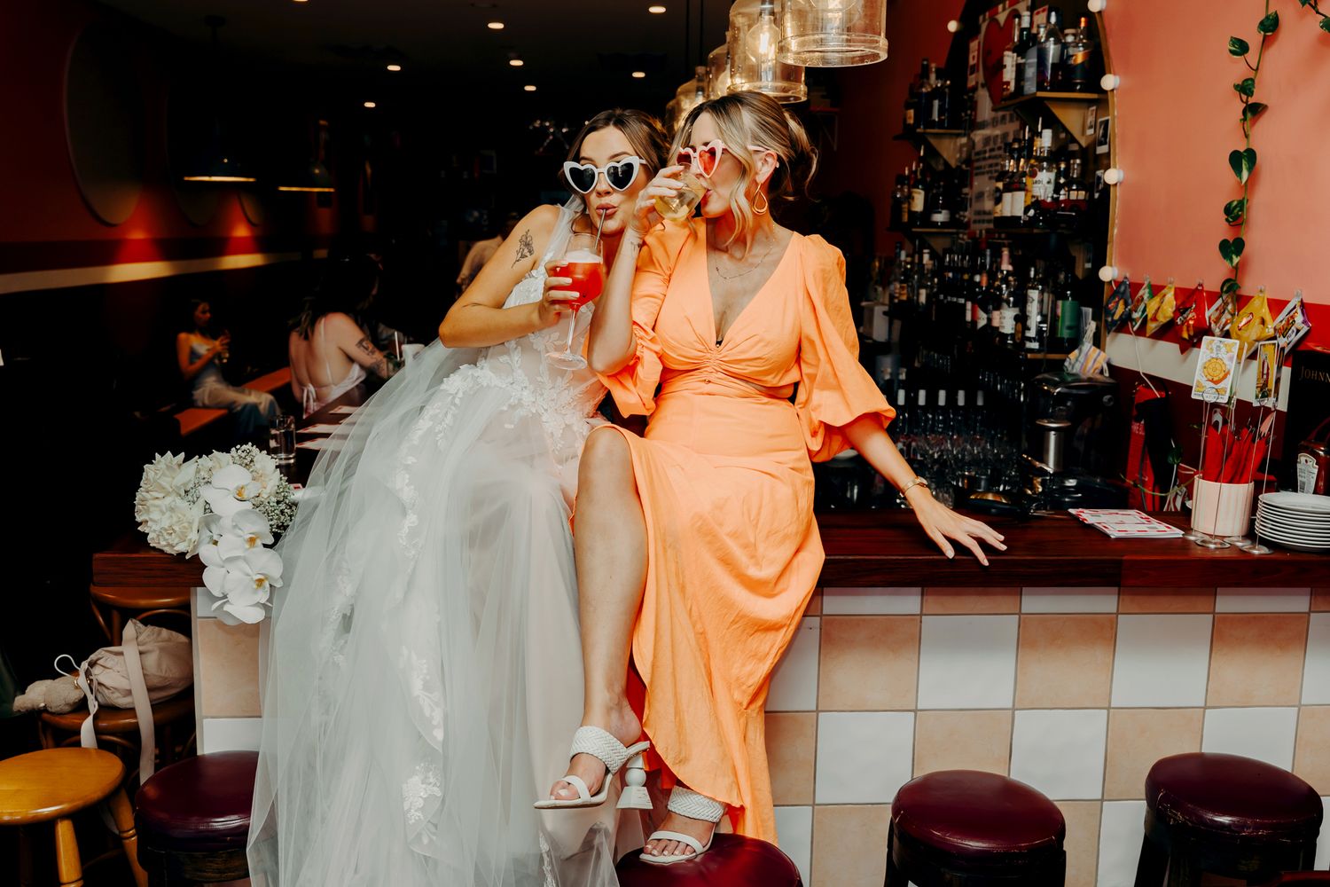 Two people in formal attire share drinks at a retro-style bar with checkered counter and warm lighting.