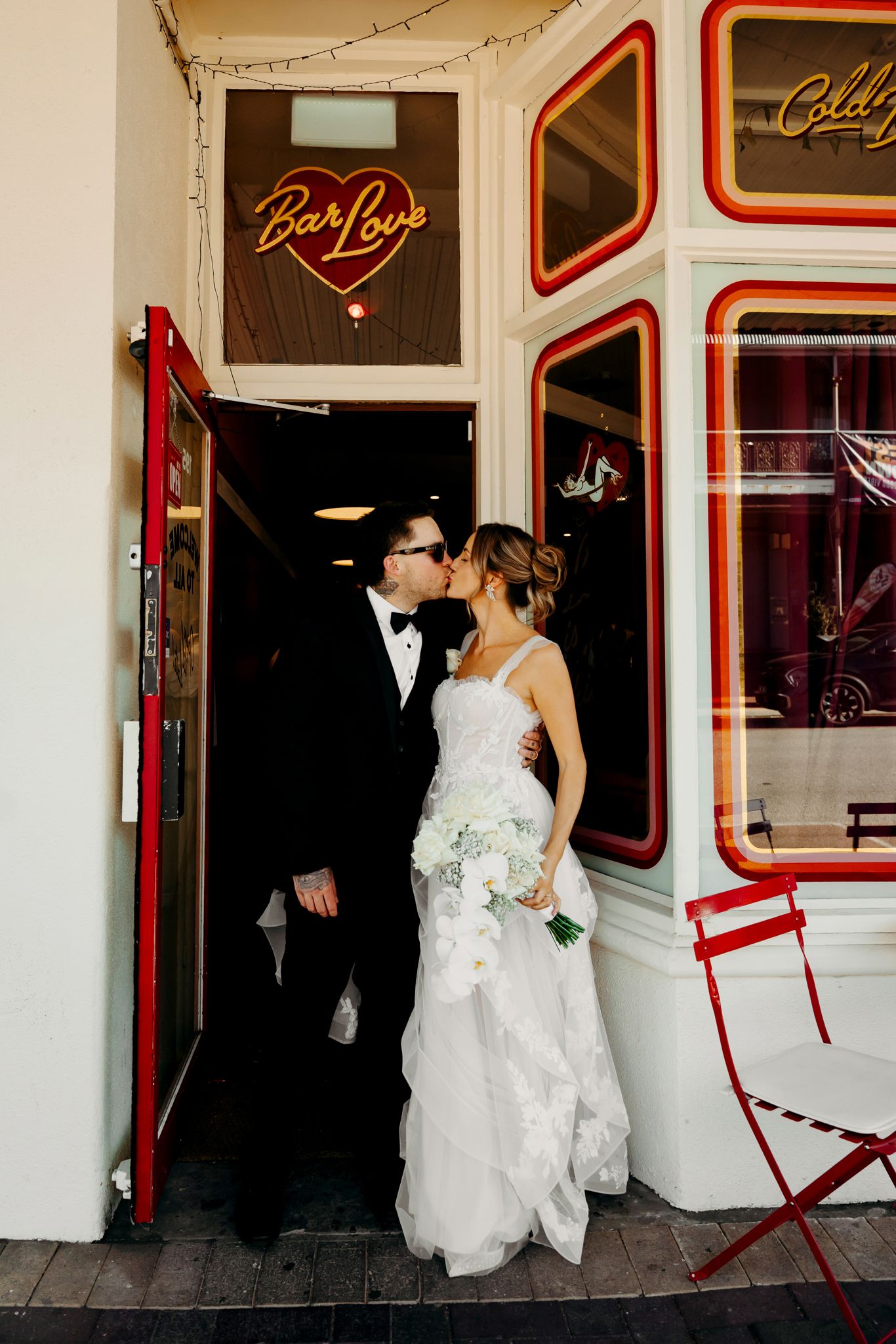A wedding couple poses in front of a white building with red accent trim and string lights.