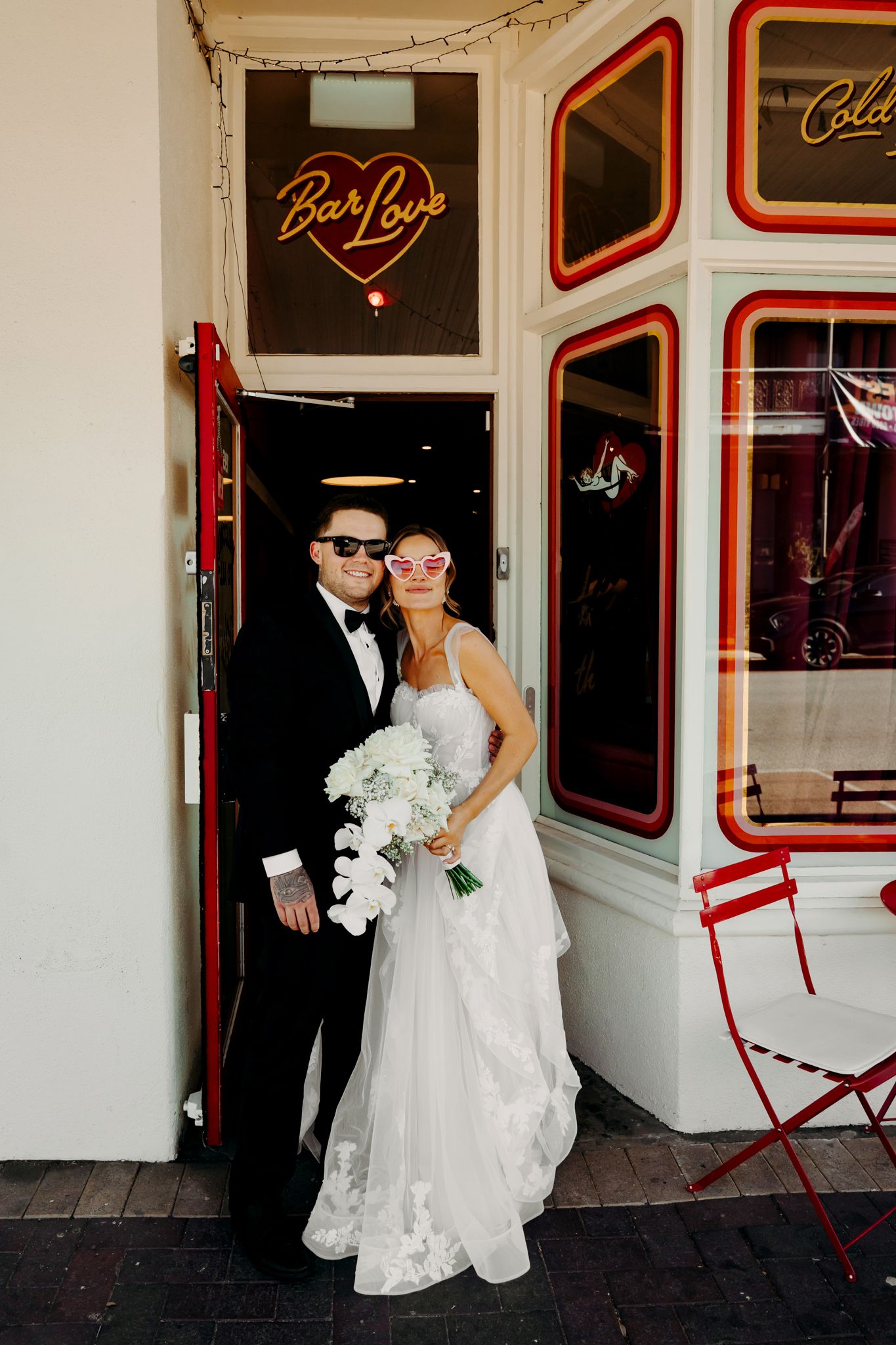 A wedding couple stands outside a vintage-style storefront with red trim and white walls.