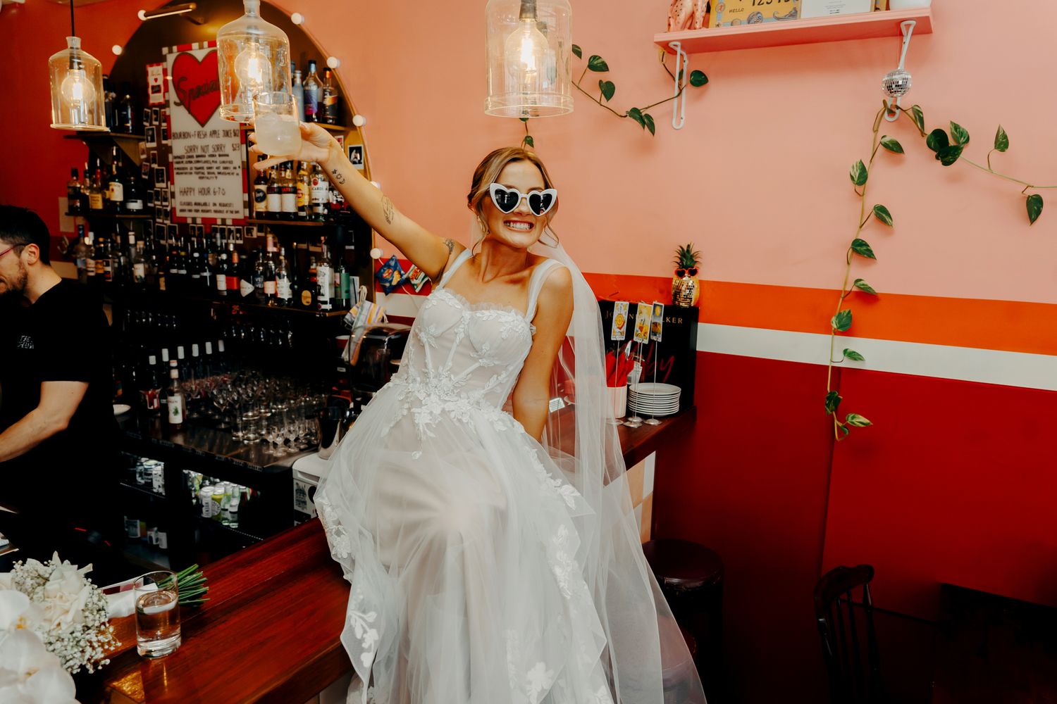 A person in a white wedding dress and sunglasses sits casually at a bar with red walls and hanging plants.
