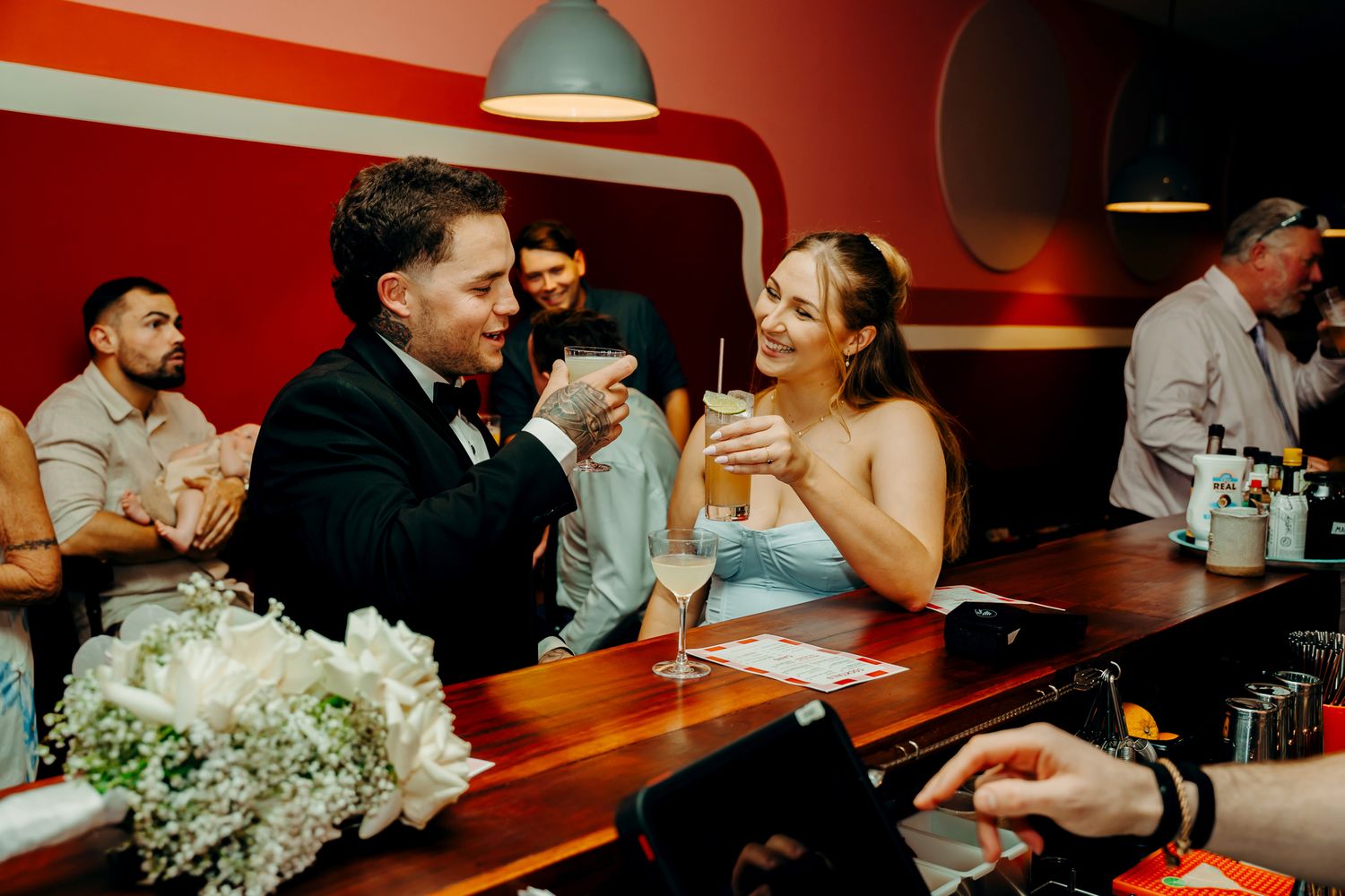 Wedding celebration scenes in a bar with red walls and pendant lighting showing candid moments between guests.