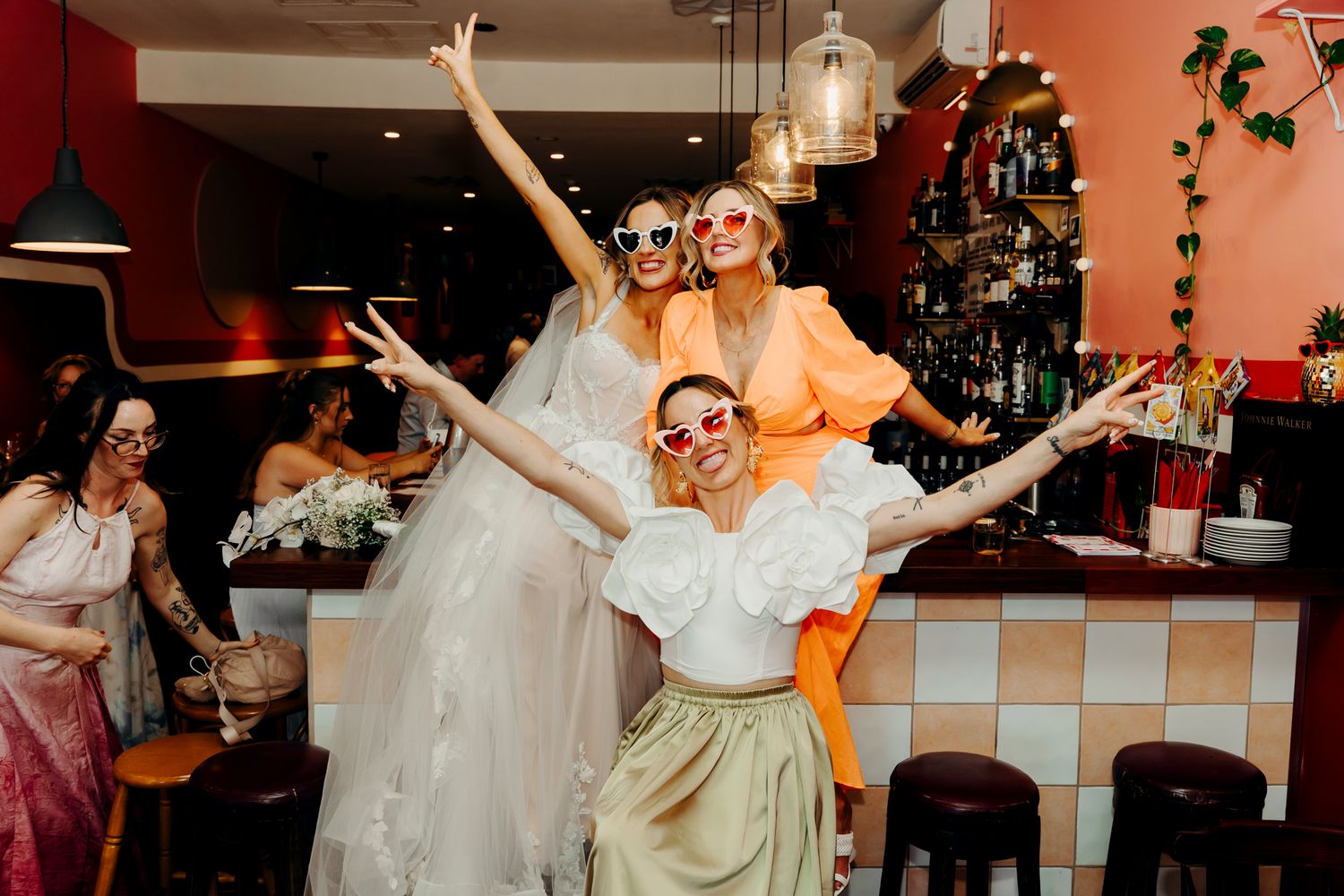 Three friends celebrating with joyful poses and raised arms at a festive party with decorative lights and greenery.