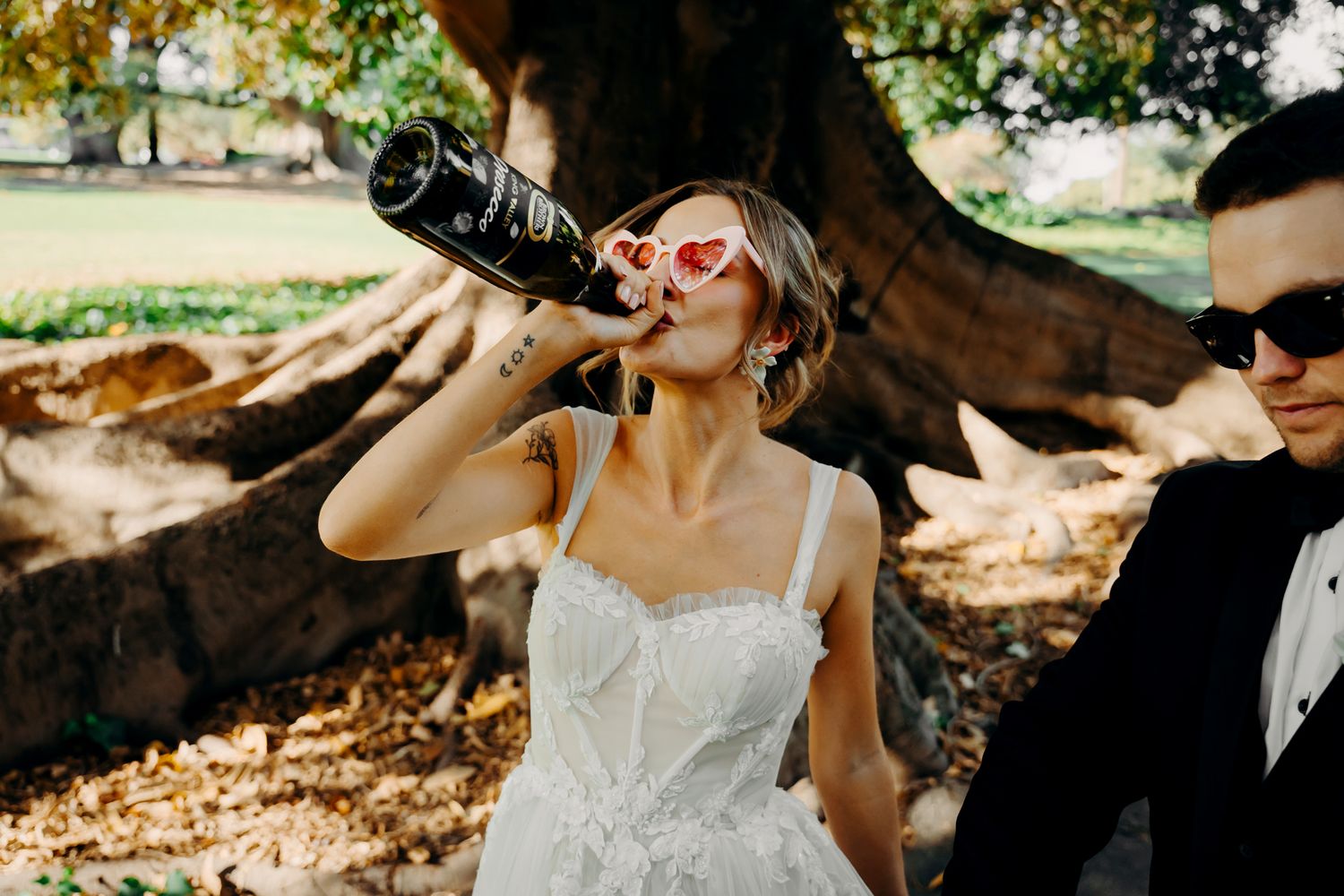 A couple in formal wedding attire shares a drink through straws in a garden setting.