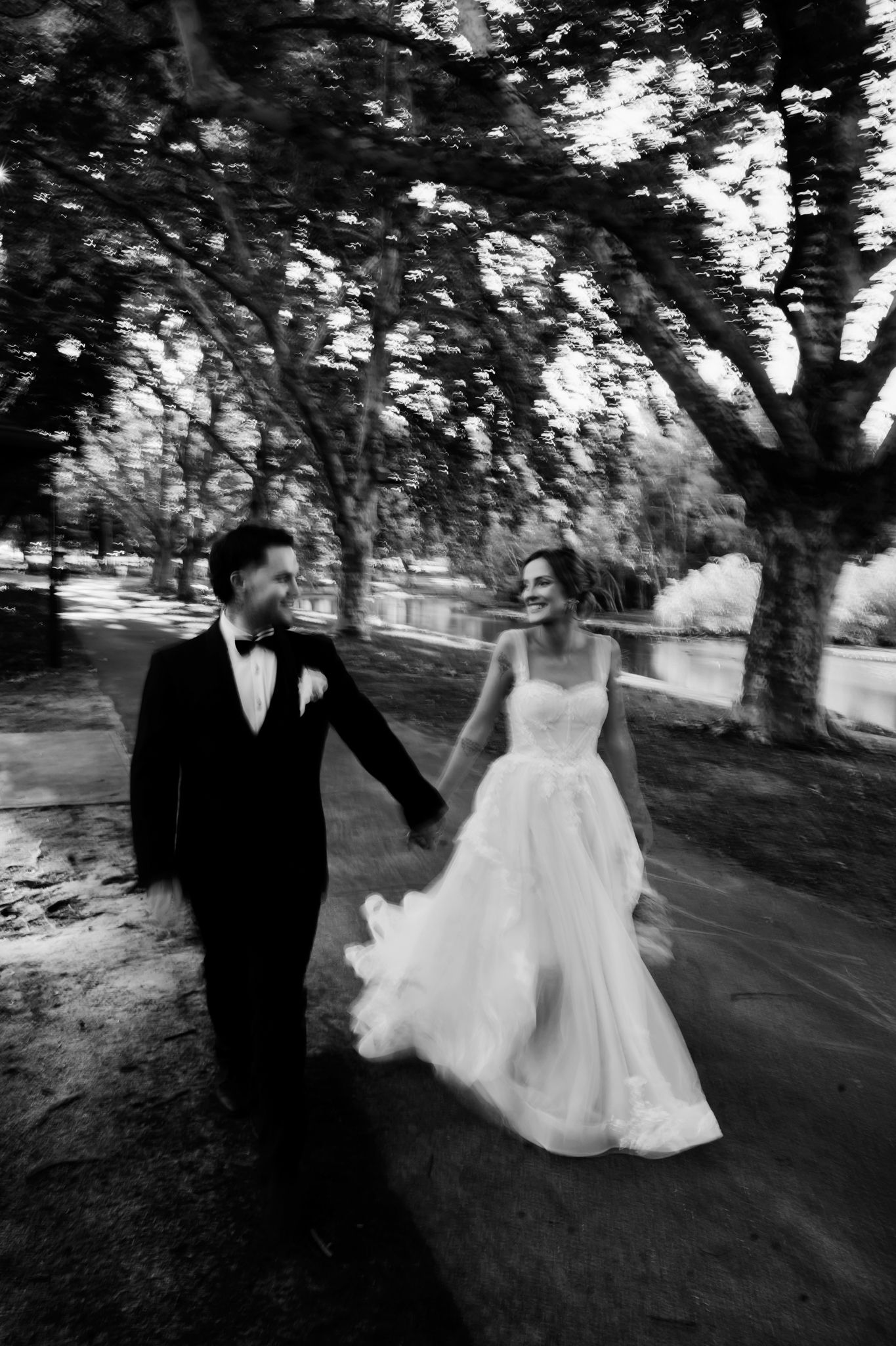 A romantic black and white wedding photo of a couple walking together under trees at an outdoor venue.