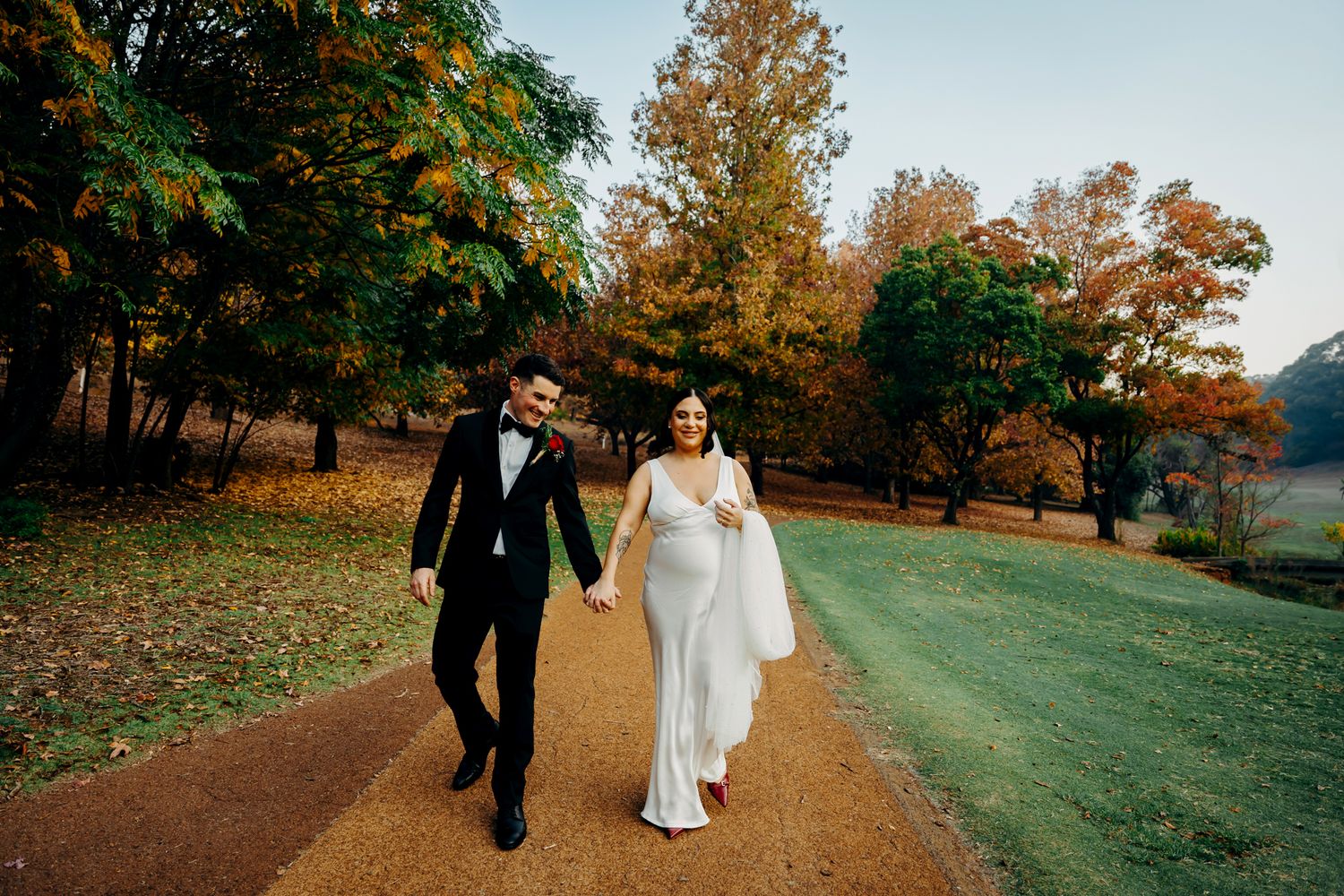 A couple walks hand in hand on an autumn path surrounded by golden trees and fallen leaves on their wedding day.