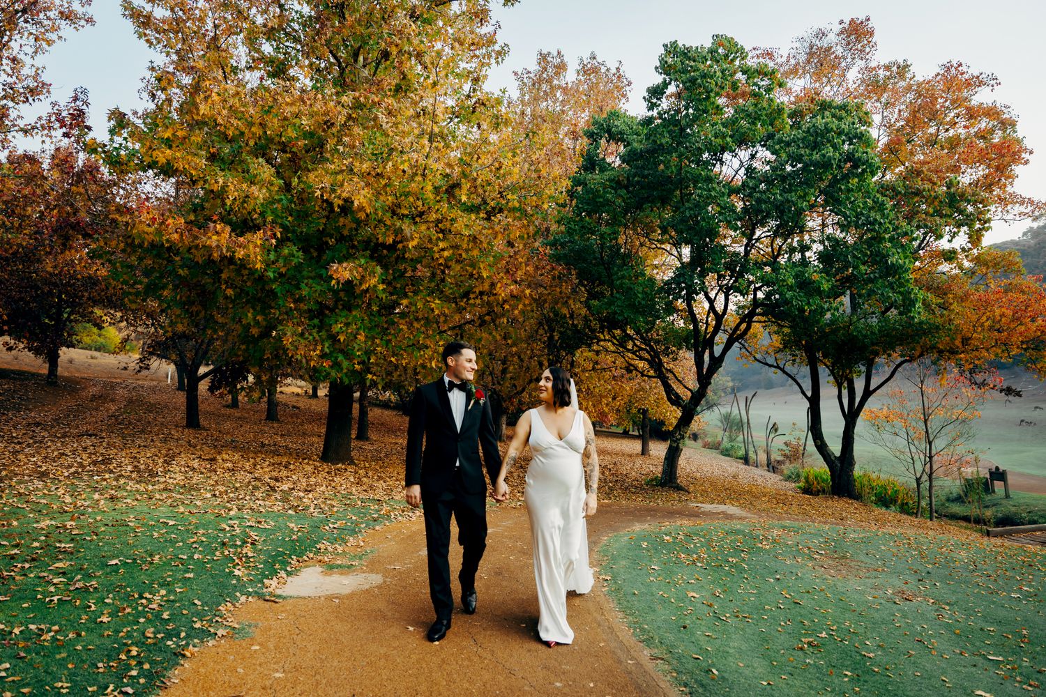 A couple walks hand in hand along an autumn path bordered by trees with colorful orange and green foliage.