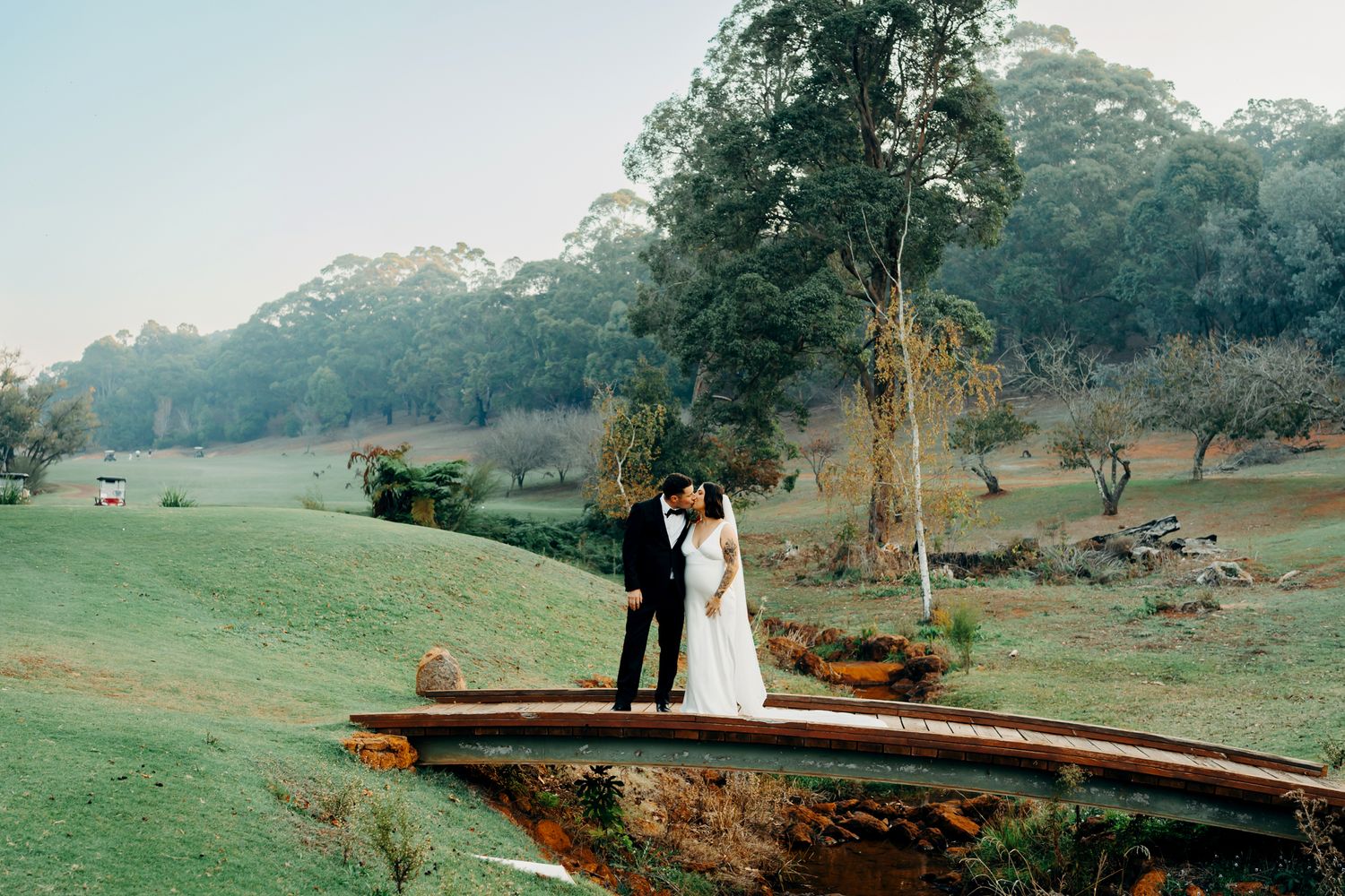 A couple poses for outdoor wedding photos in a rustic setting with green fields and a wooden log bridge.