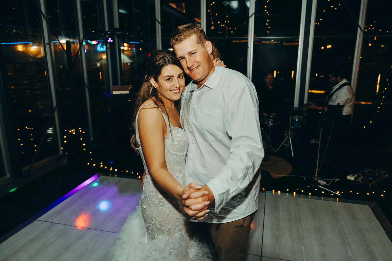 A couple dances together in an elegant venue with glowing lights and windows in the background.