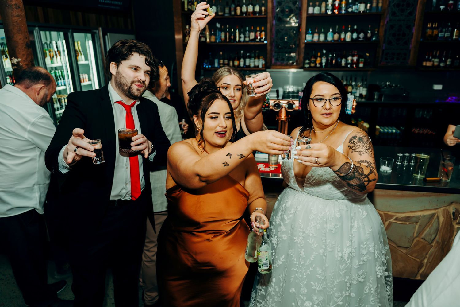 Wedding guests raise their glasses in celebration during a reception at a bar venue with rustic decor and warm lighting.