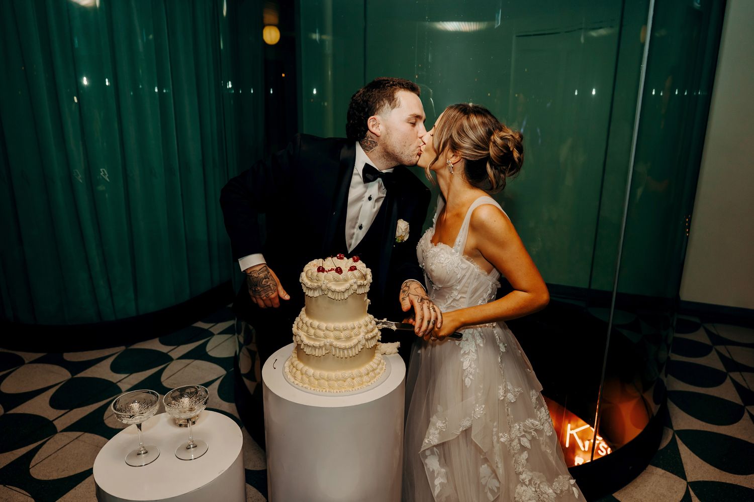 Newlyweds share a romantic kiss while cutting their elegant three-tiered wedding cake at an intimate reception.