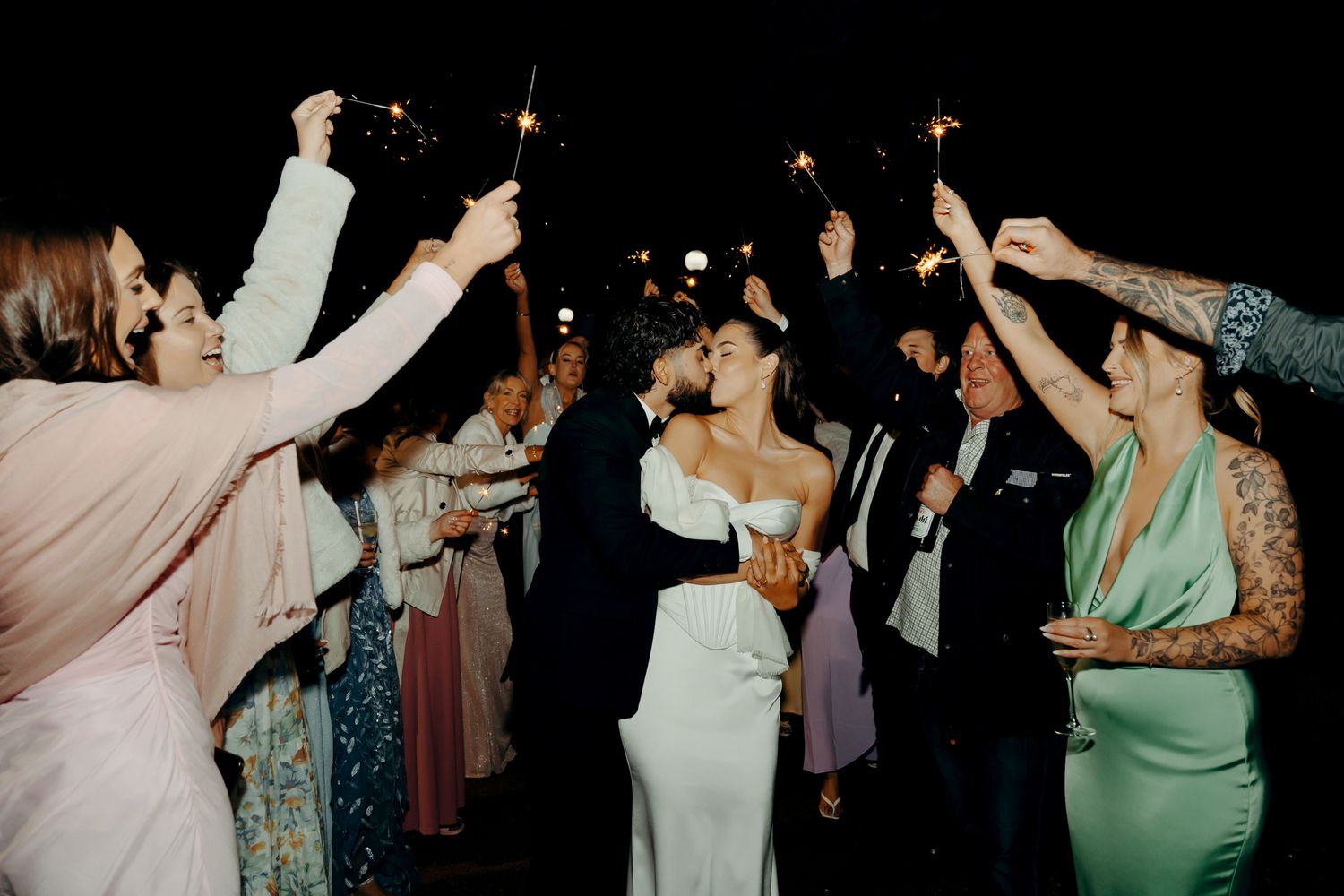 Newlyweds share a romantic kiss surrounded by guests holding sparklers during a joyful outdoor wedding reception at night.