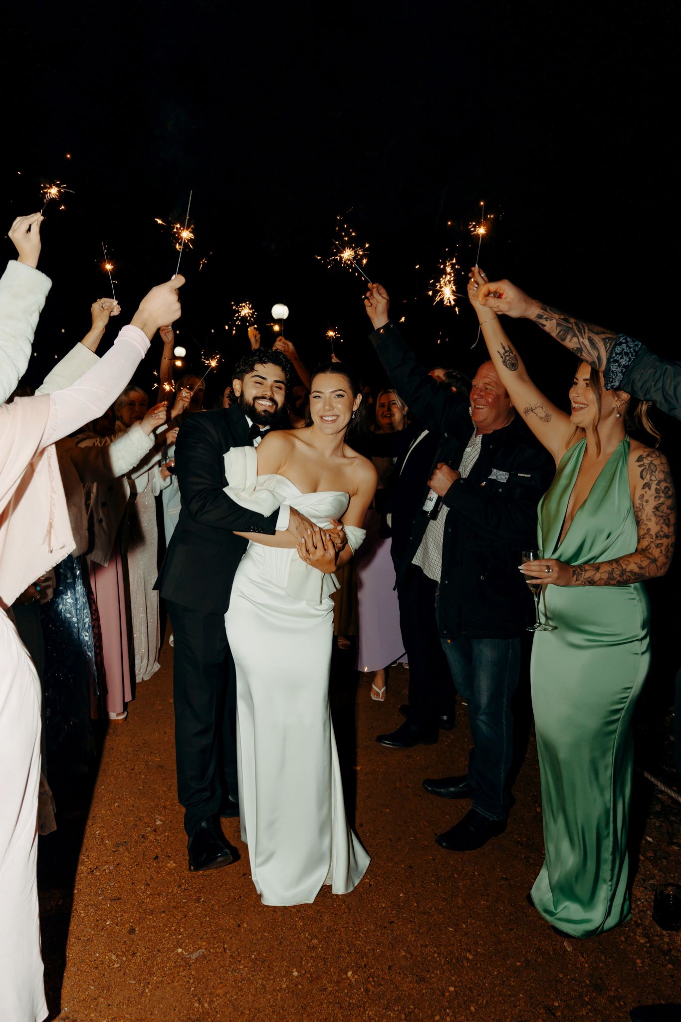 Bride and groom walk through sparkler send-off with smiling wedding guests at night outdoor celebration.