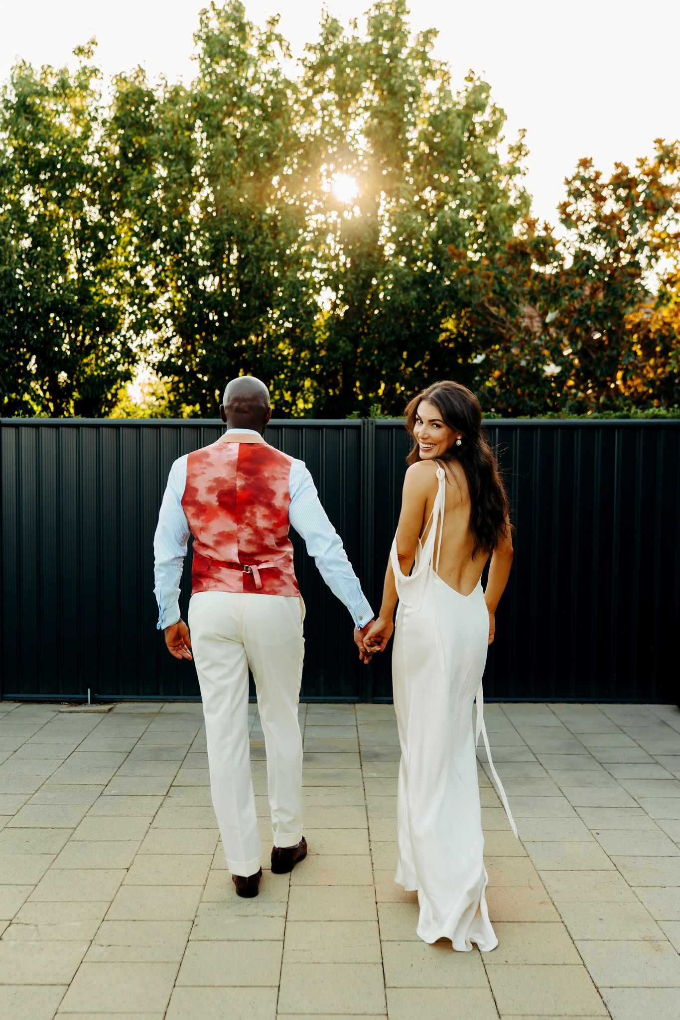 A bride in a backless white gown and groom in a red vest hold hands outdoors at sunset.
