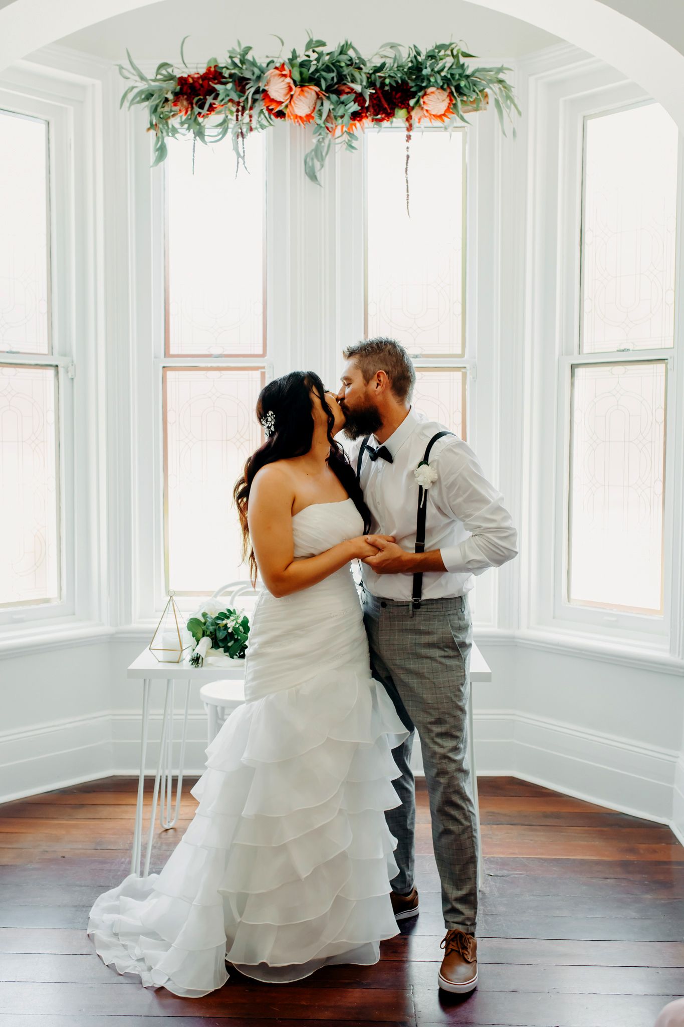 Couple sharing an intimate moment under floral arch decor in bright white bay window setting.