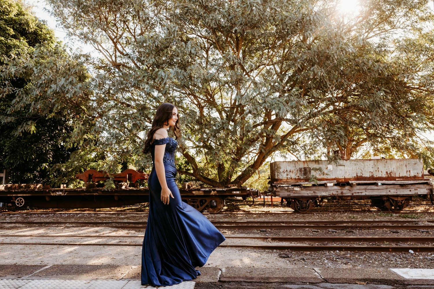 Woman in elegant white dress standing on train platform with vintage carriages in background.