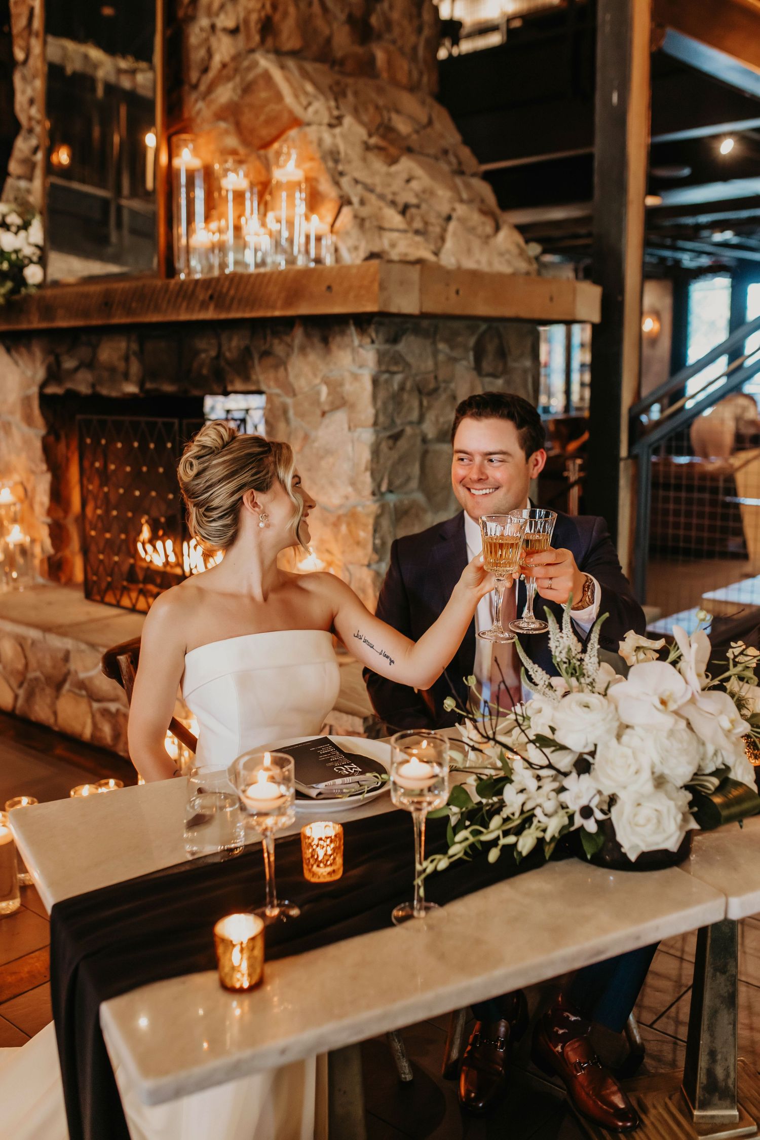 Wedding couple toasts at rustic reception with stone fireplace and candlelight.