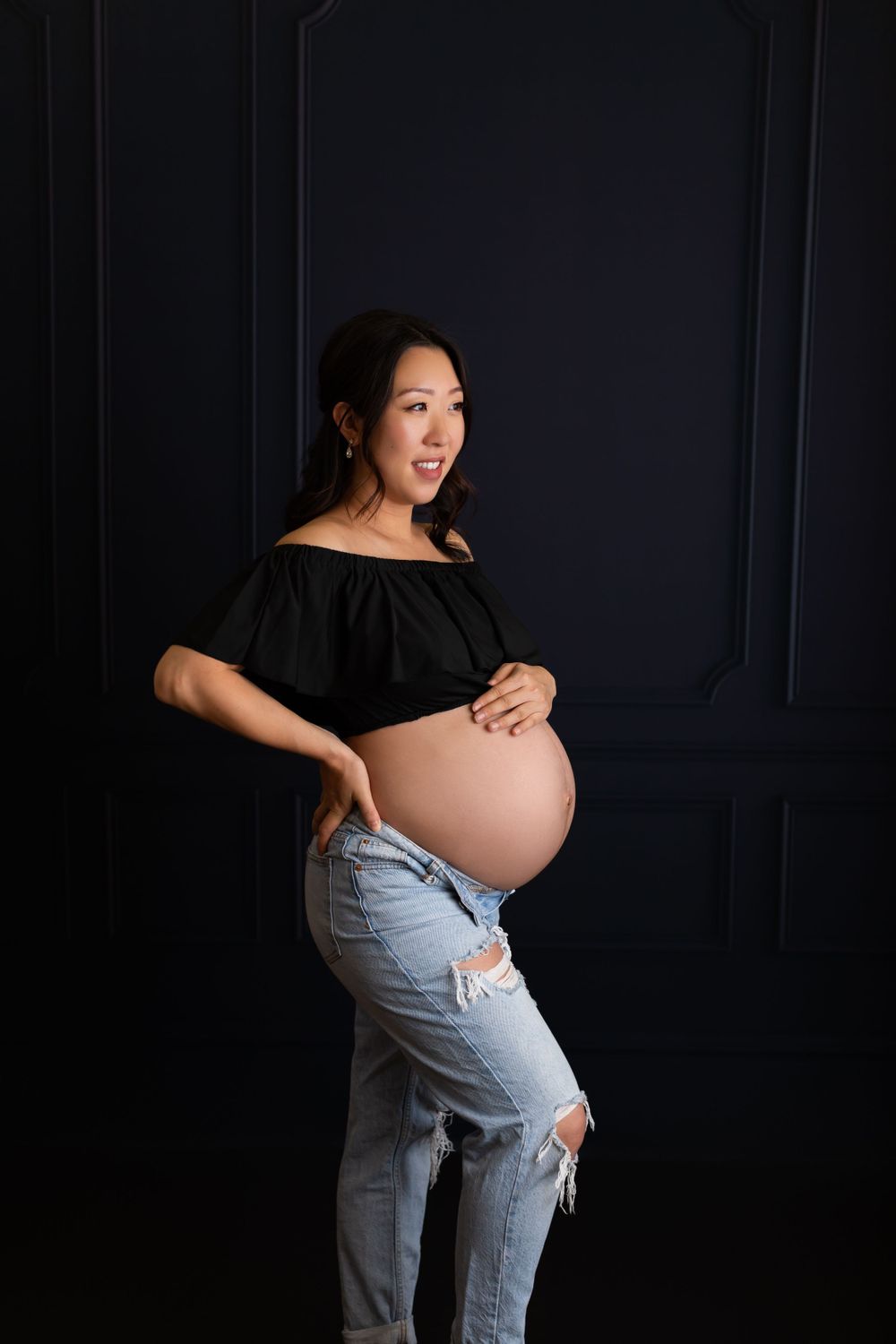 A maternity portrait against a dark wall shows an expectant mother in a black off-shoulder top and distressed jeans.