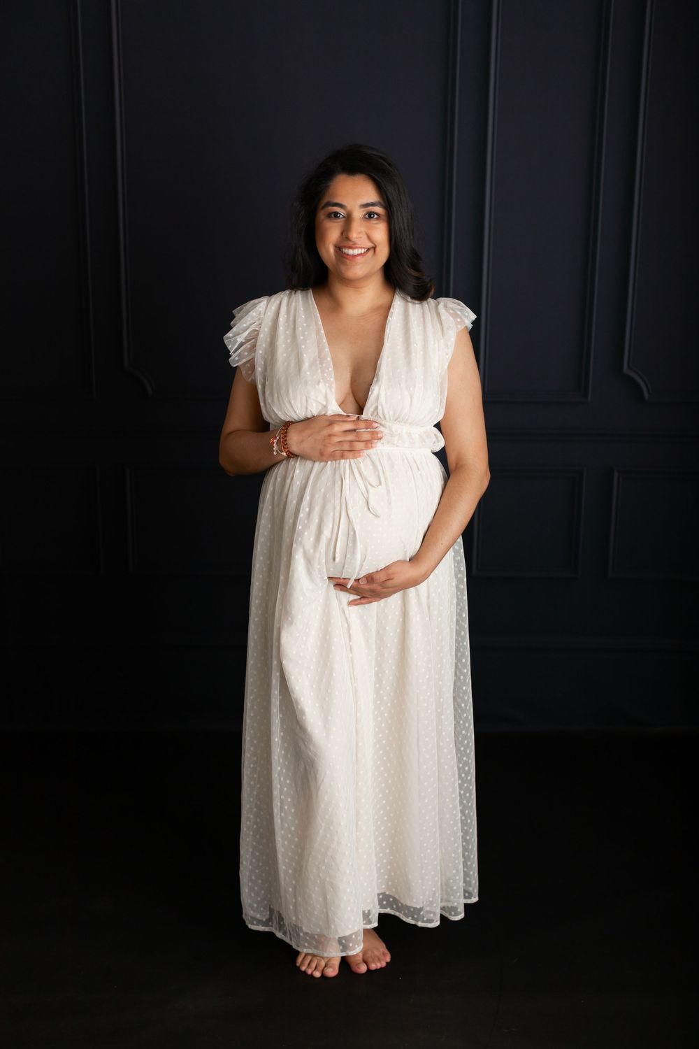 Expectant person in white lace dress poses against dark wall during maternity photoshoot.