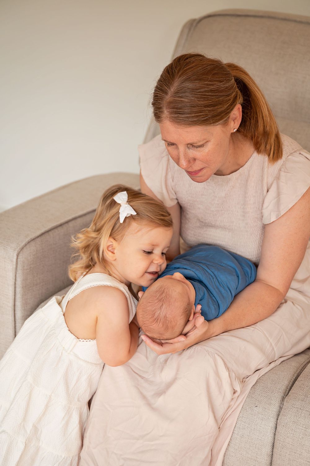 A tender family moment on a couch as a young child in a white dress leans over to look at a baby.