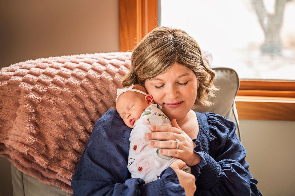 A tender moment between parent and newborn baby in a cozy chair by a window with soft natural light streaming in.
