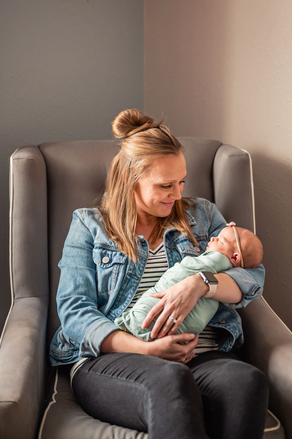 A person in a denim jacket sits in a gray chair holding and gazing lovingly at a newborn baby.