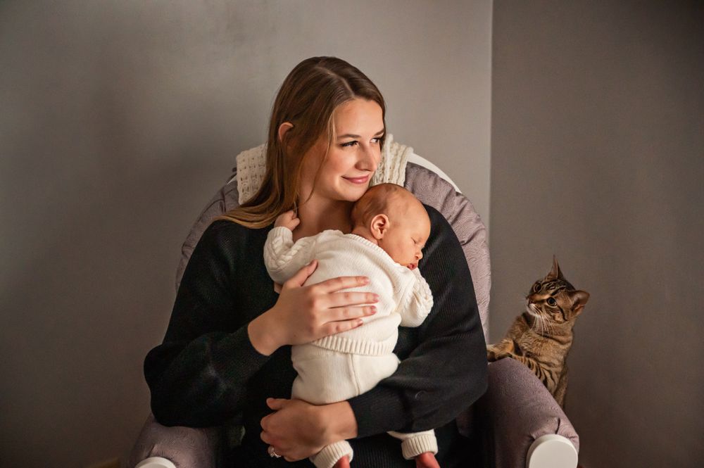 Tender moment between parent and infant with cat sitting nearby in grey room.