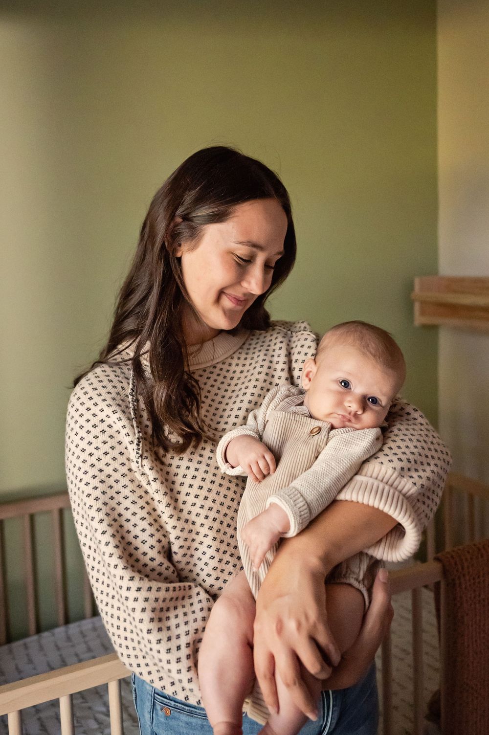A tender moment between parent and baby in a nursery, wearing a polka dot sweater and holding the infant near a crib.
