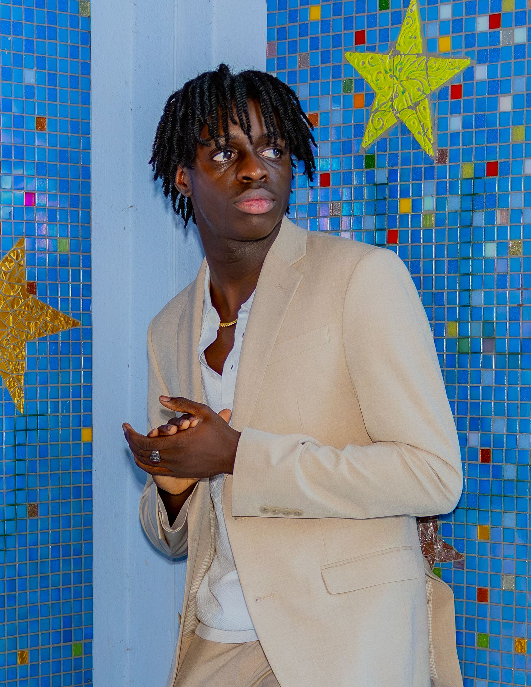 A person in cream blazer poses against a blue mosaic wall decorated with golden stars.
