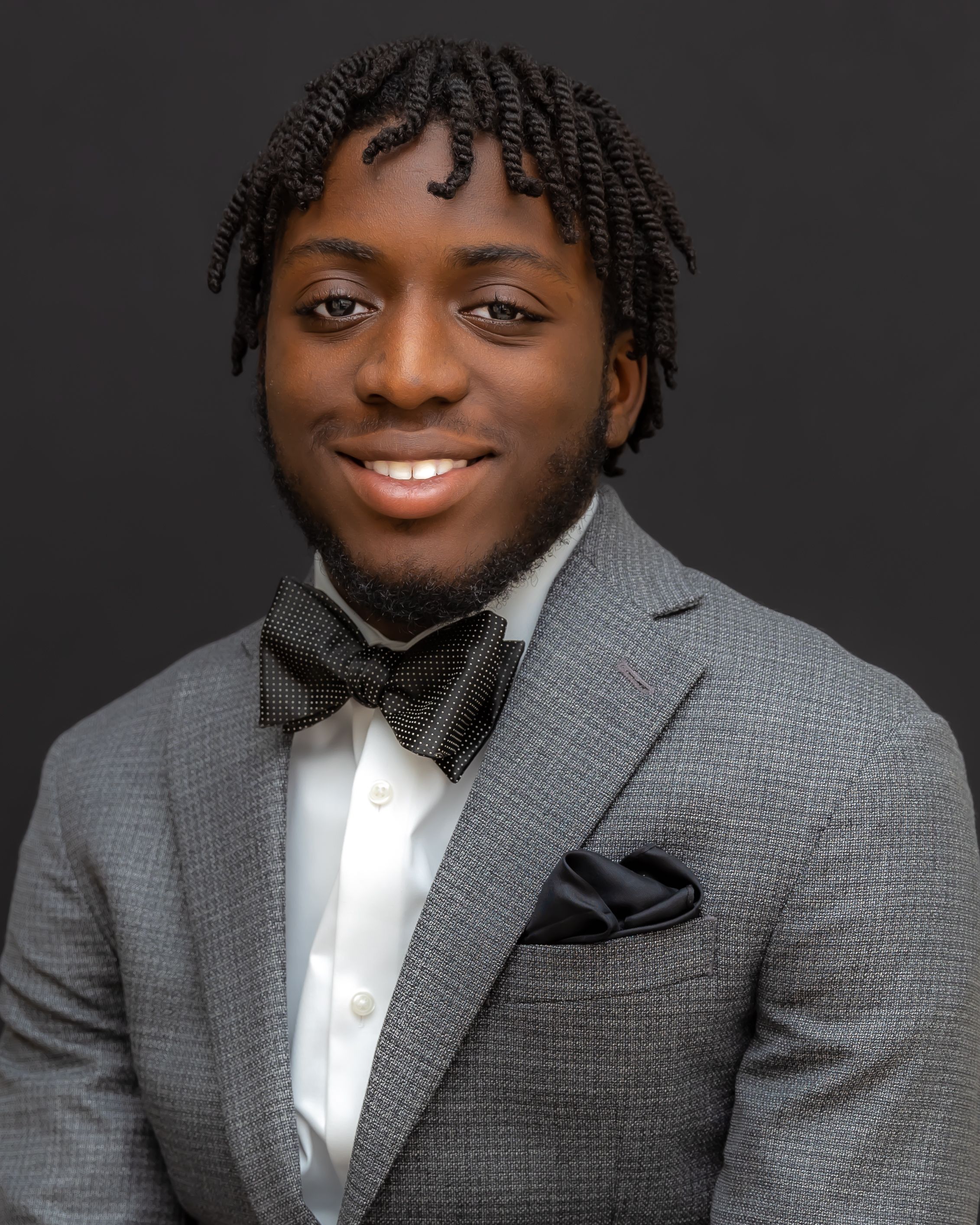 A professional headshot of someone in a grey suit with black bow tie and textured hairstyle.