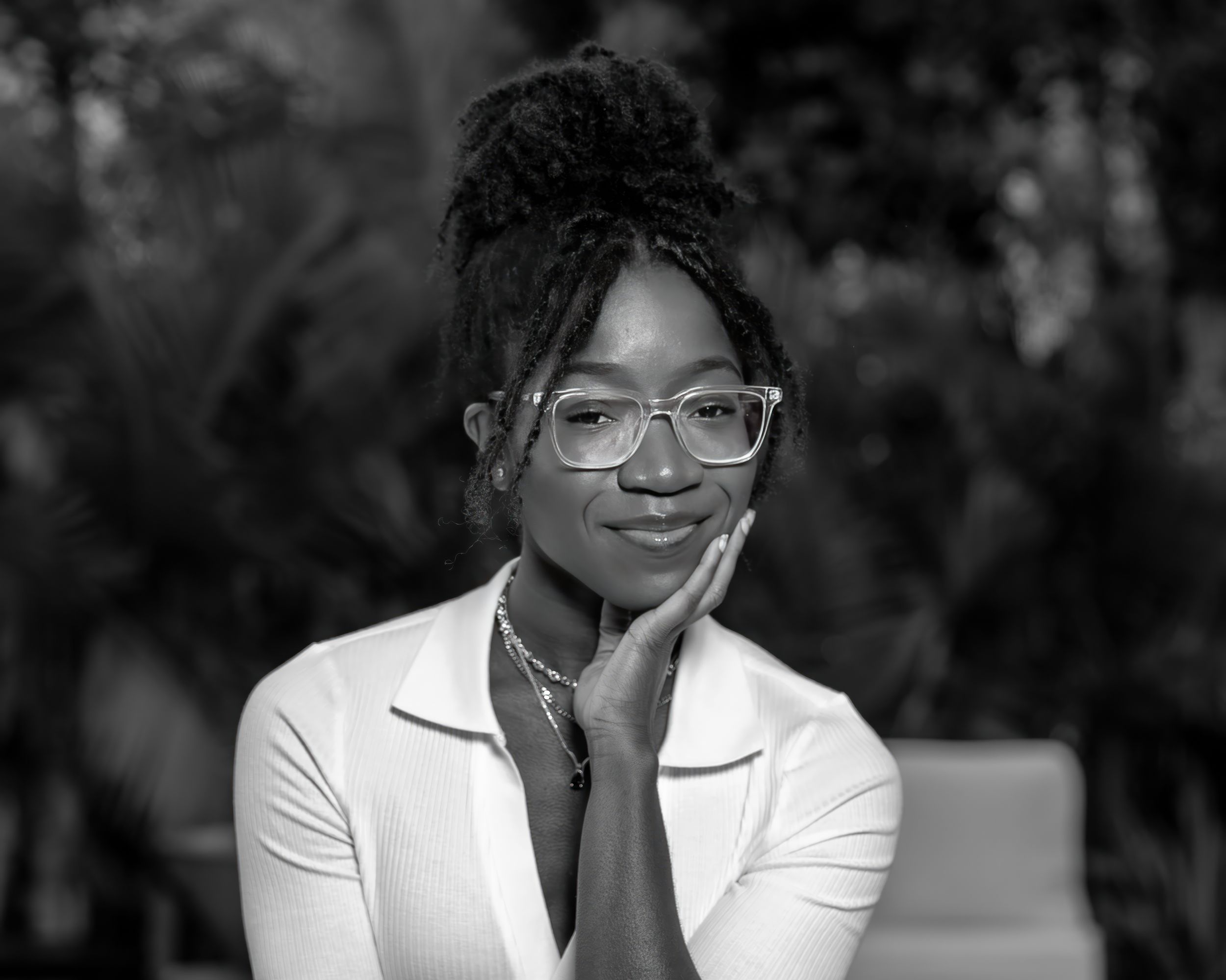 A black and white professional portrait of someone in glasses and white blazer with a contemplative expression.