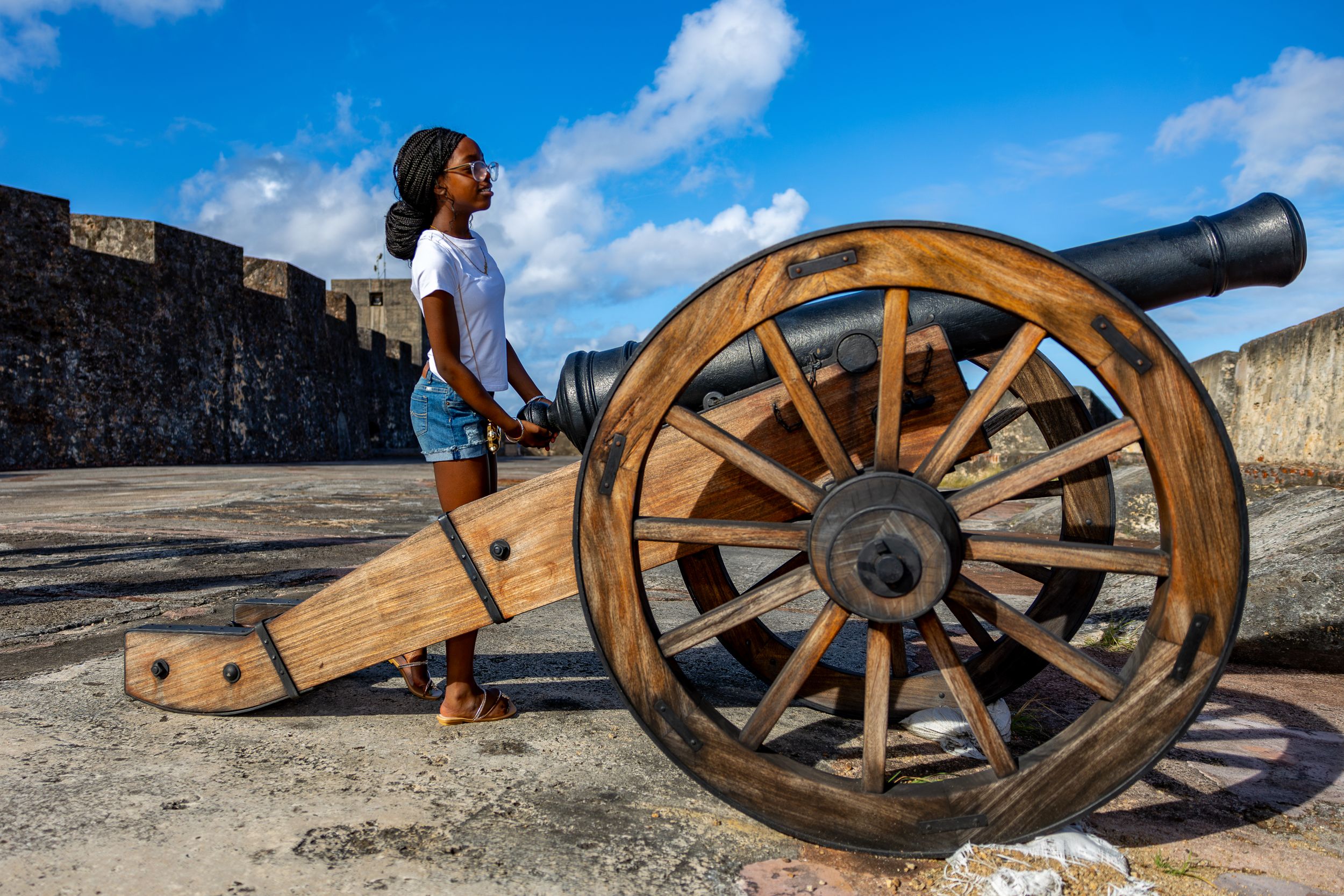 An old cannon sits on a historic stone fortress wall against a blue sky with white clouds.