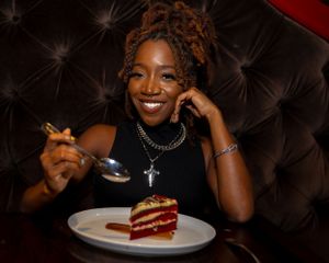 Someone enjoying a slice of red velvet cake at a restaurant with dark tufted seating.