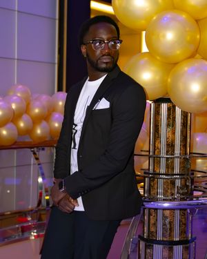 Formal event with someone in a black blazer standing by gold balloons and decorative columns.