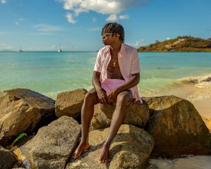 A person sits contemplatively on coastal rocks overlooking a turquoise Caribbean beach.