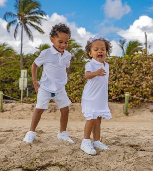 Two children in white outfits playing and laughing on tropical beach with palm trees.