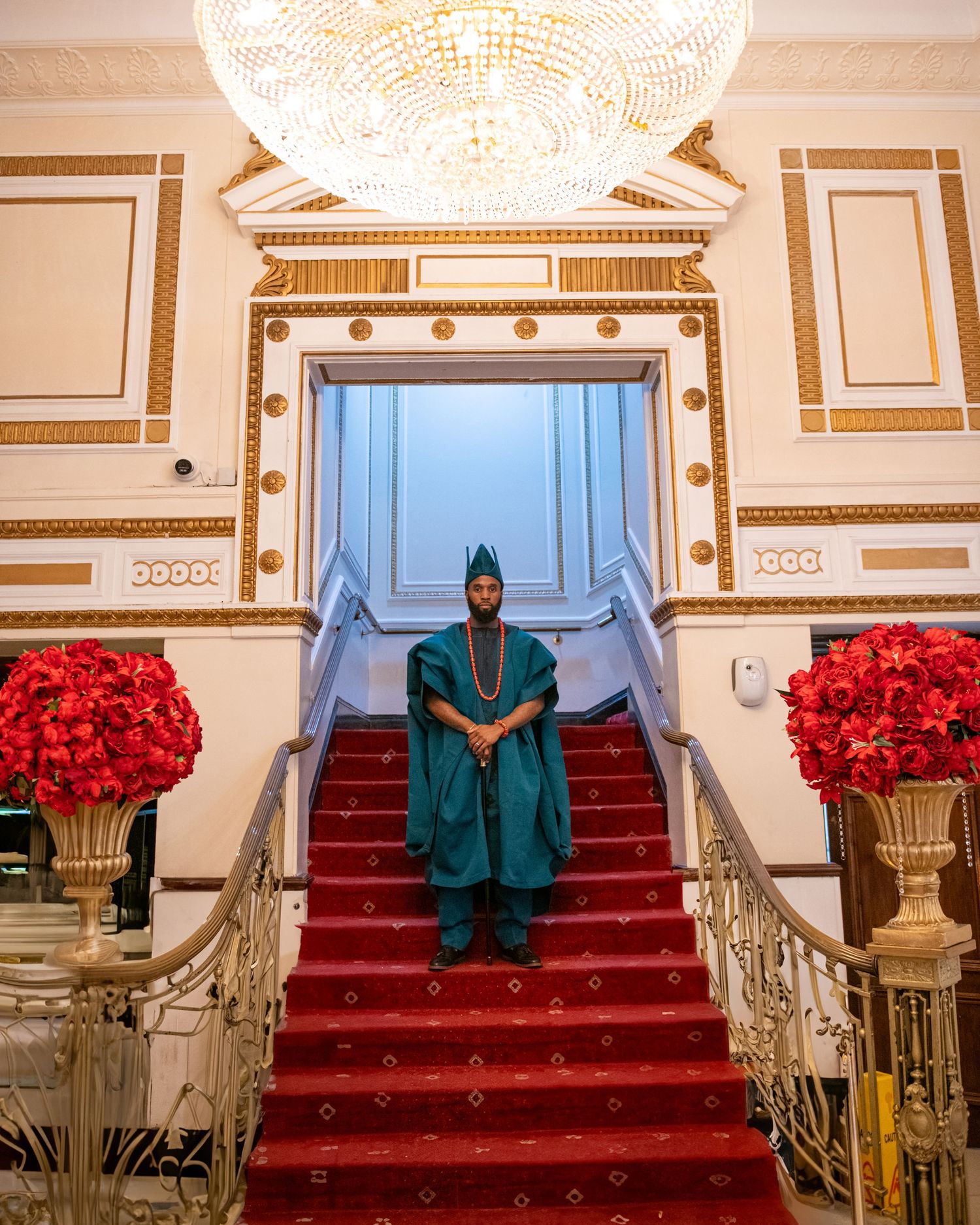 Man in teal Nigerian traditional agbada outfit stands on red-carpeted staircase in ornate gold venue.