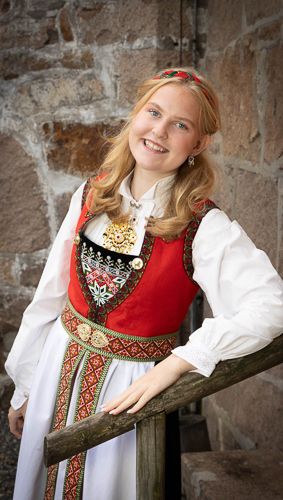Traditional Norwegian folk costume with red embroidered vest and white blouse shown in three poses against stone wall.