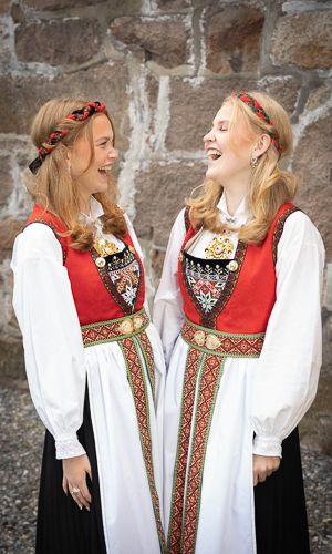 Two people wearing traditional Norwegian folk costumes called bunads with red vests and white blouses share a joyful moment.