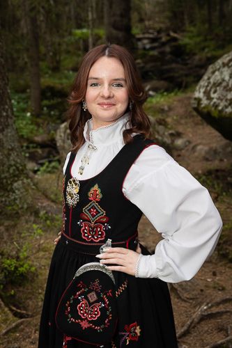 Traditional Norwegian folk costume with black embroidered vest and white puffy sleeves shown in forest setting.