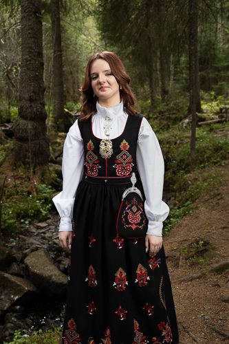 Traditional Norwegian folk costume with black embroidered dress and white blouse photographed in a forest setting.