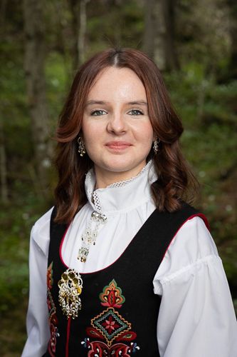 Traditional Norwegian bunad dress with embroidered black vest, white high-neck blouse, and ornate silver brooch against forest backdrop.